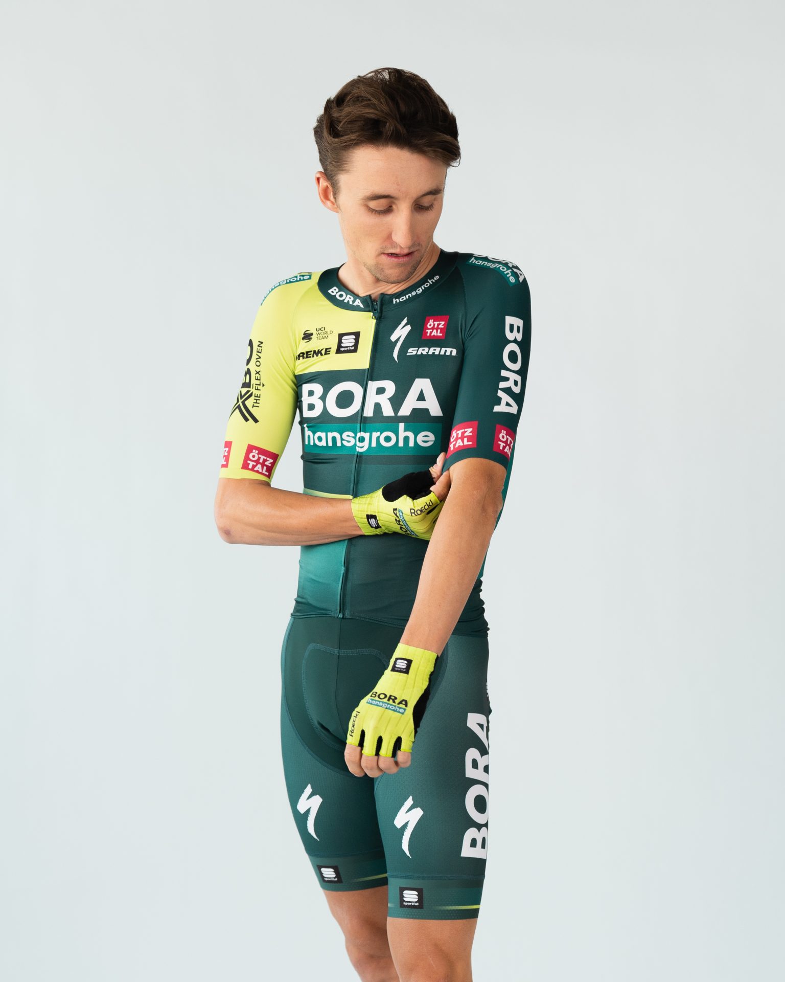Jai Hindley models the new Bora-Hansgrohe team kit for 2024. It's mostly forest green, with bright blocks of lime on the right shoulder and gloves. Jai is looking down as he adjusts a sleeve, with a neutral expression on his face.