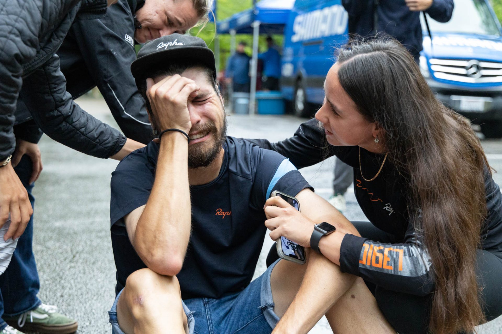 Sam Boardman sits on the pavement and briefly cries after winning stage 4 of the Joe Martin Stage Race. It's well after the stage, and he's dressed in street clothes - jean shorts, a Rapha t-shirt and cap, and he has one hand to his face with his eyes closed as he's overcome with emotion.