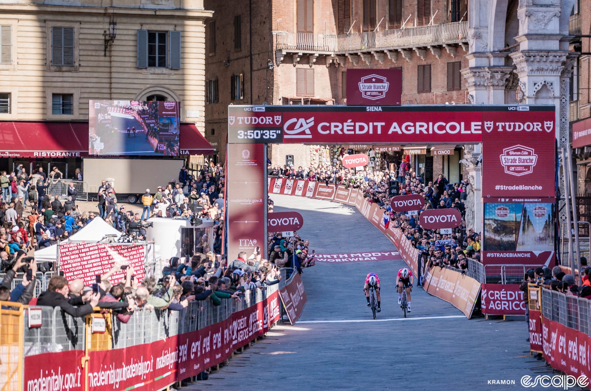 Lotte Kopecky and Demi Vollering are in a photo finish on the line in the Campo square in Siena at Strade Bianche, thronged by fans as both throw their bikes at the line.