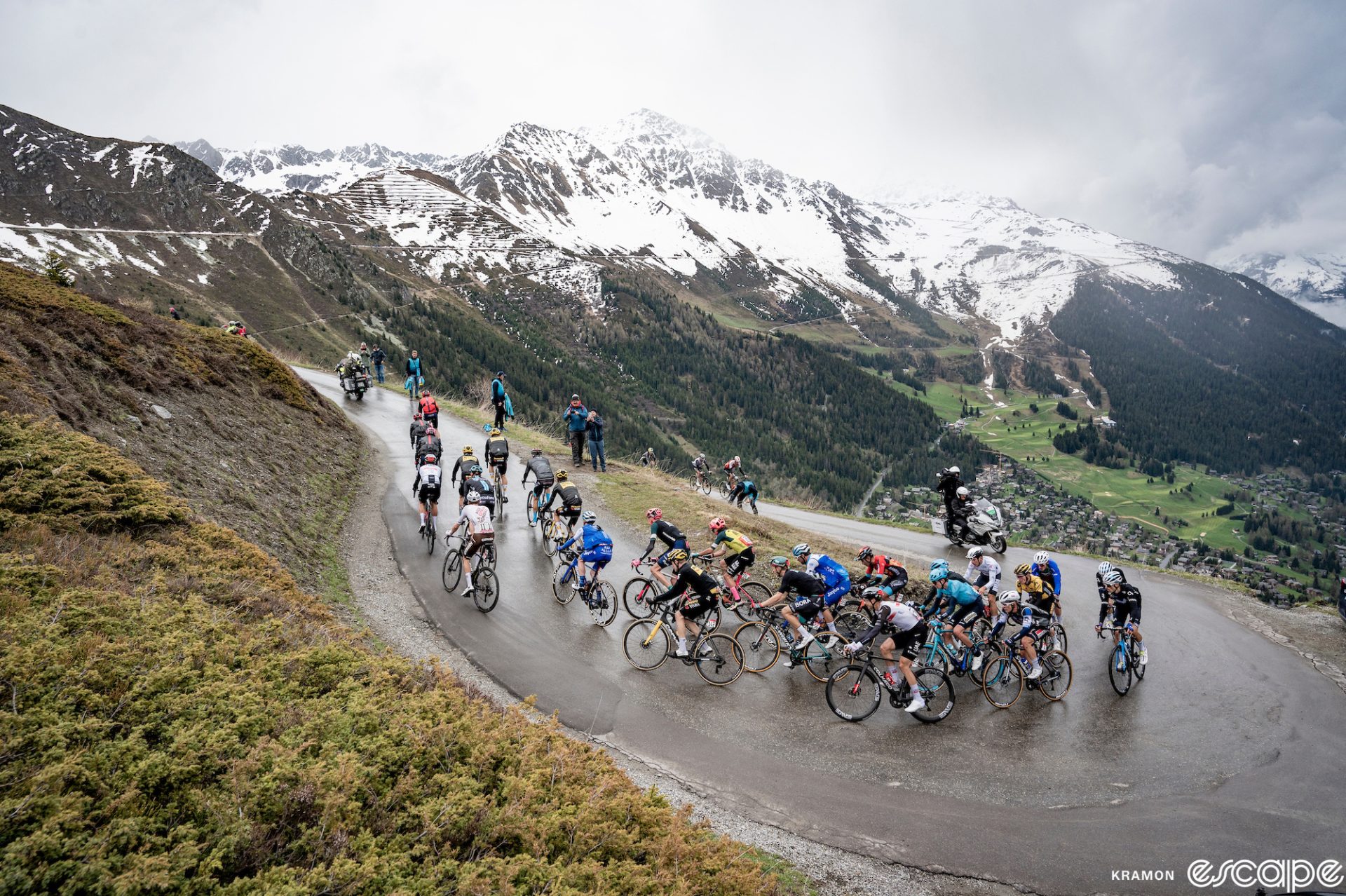 A cold, wet pack of riders climbs a switchback on the Croix de Coeur climb in the Giro d'Italia. Grey skies and snowy mountains loom beyond.