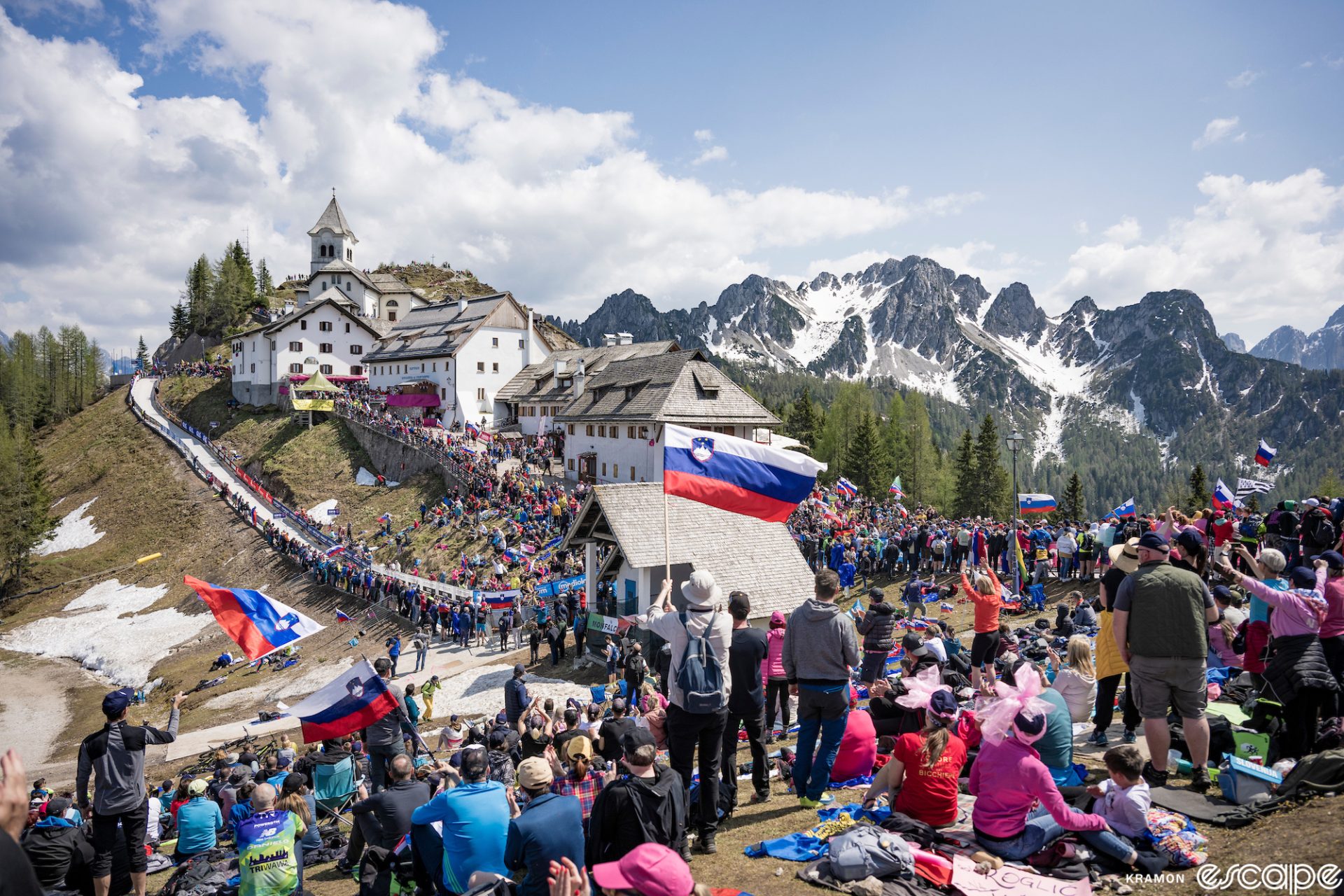Crowds, many waving Slovenian flags, pack the slopes of the Monte Lussari monastery on stage 20 of the Giro d'Italia. Rugged, glaciated peaks loom behind as the monastery buildings are packed onto a sheer slope. 