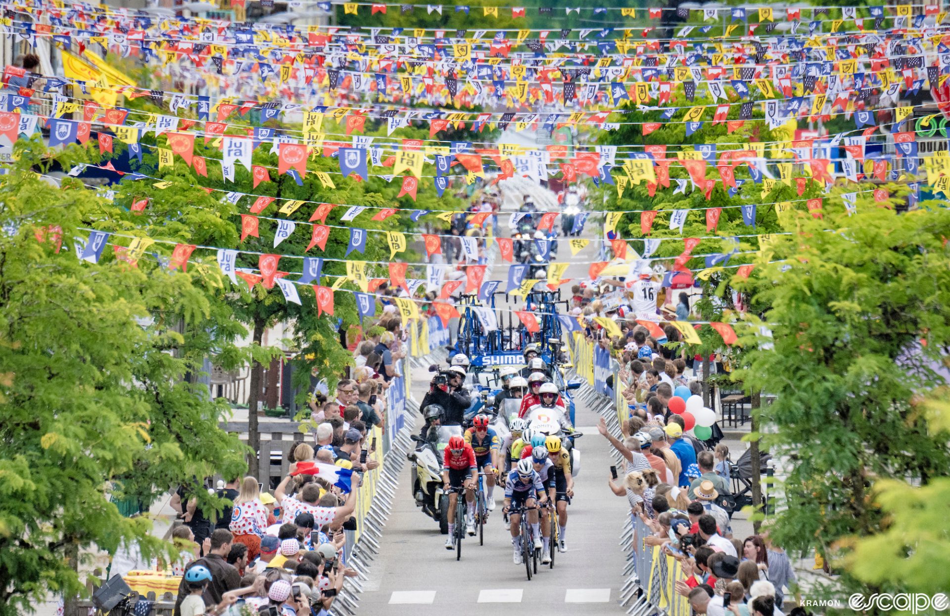 The breakaway rides through a village in the Tour de France; overhead, strings of colorful flags crisscross the road above the riders, almost like Tibetan prayer flags.