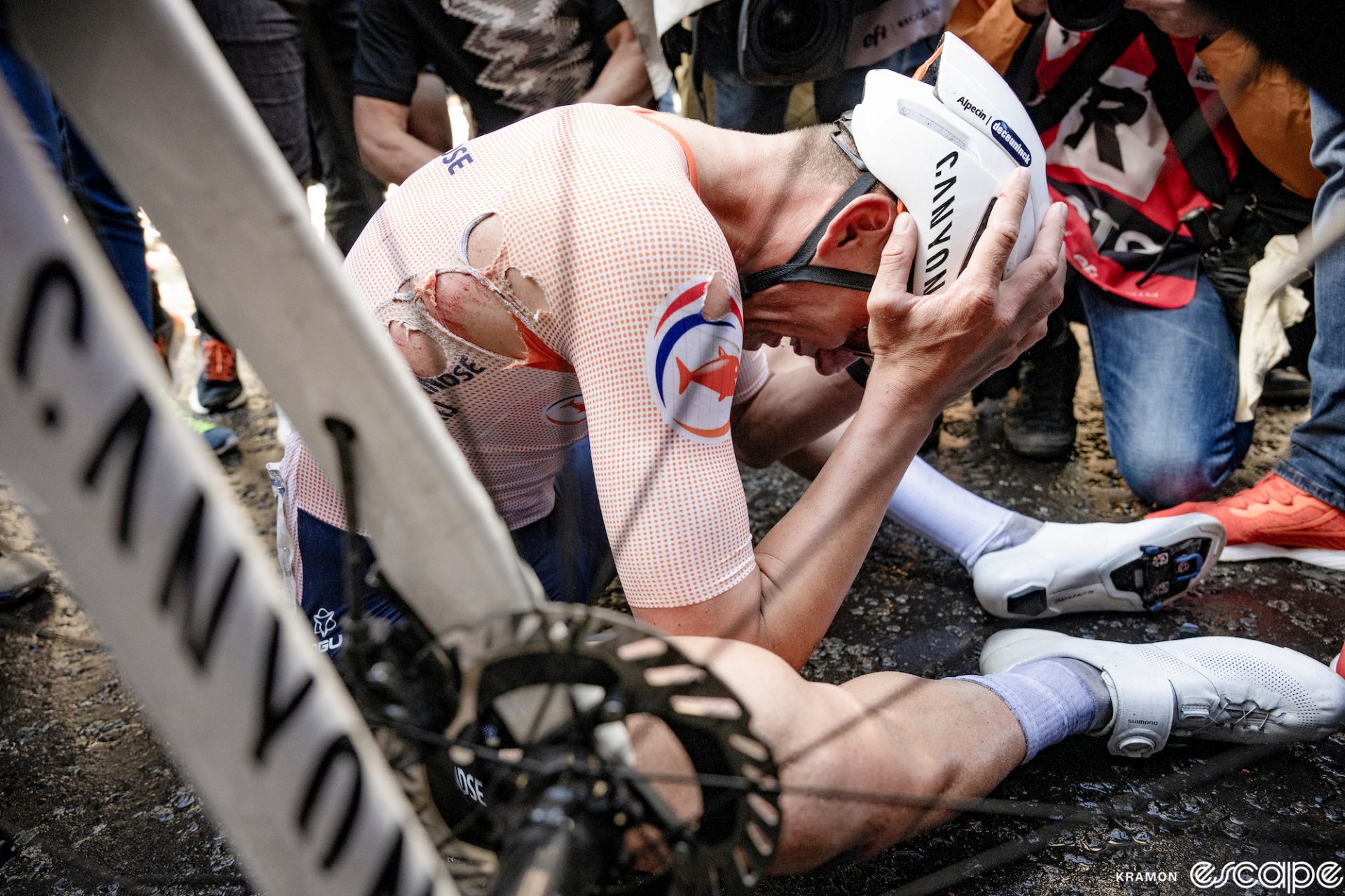 Mathieu van der Poel holds his head in his hands as he sits on the pavement after winning the World Road Championship. His bike's fork frames the picture as he's seen through the spokes of the wheel. His jersey is torn and he is wet and dirty from the racing and a late crash.