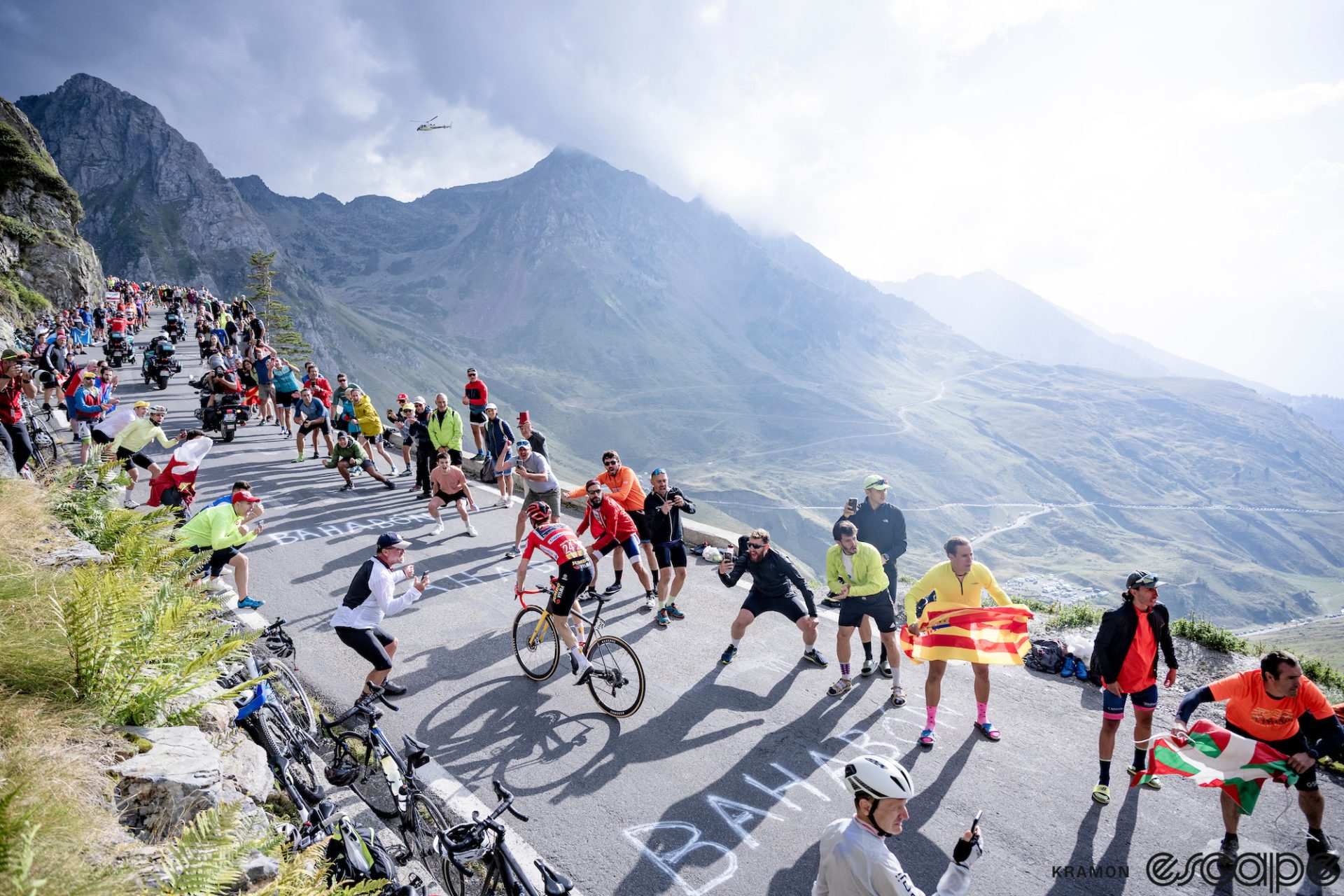 Sepp Kuss climbs the Col du Tourmalet alone in pursuit of Jonas Vingegaard. The road is lined with fans, some waving Basque and Spanish flags, and behind the Pyrenees loom in misty backlit light as a helicopter flies in the distance.