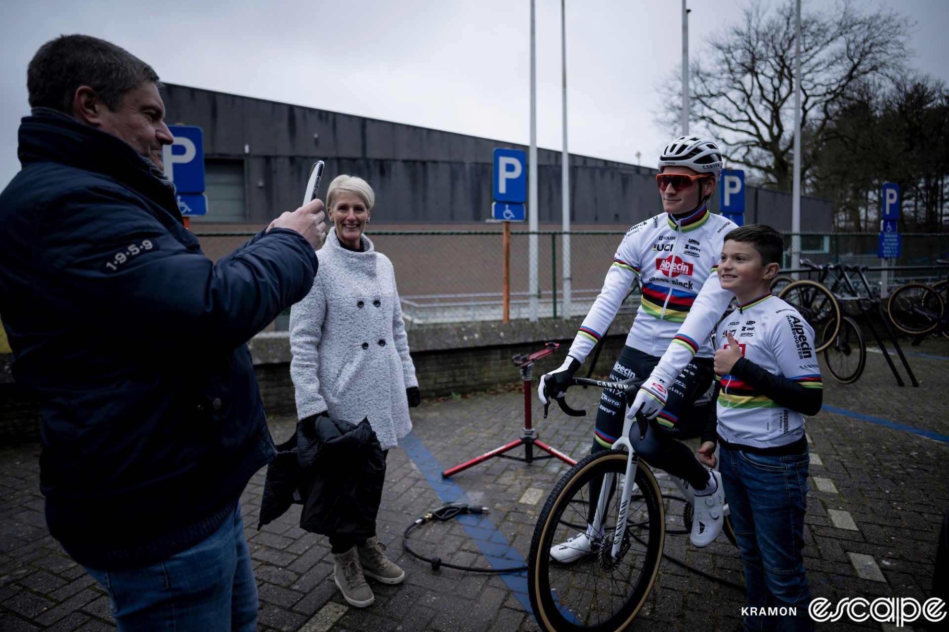 Mathieu van der Poel poses with a young fan at a cyclocross race. The young boy is smiling and giving a thumbs up for his father who is taking a cell phone photo.