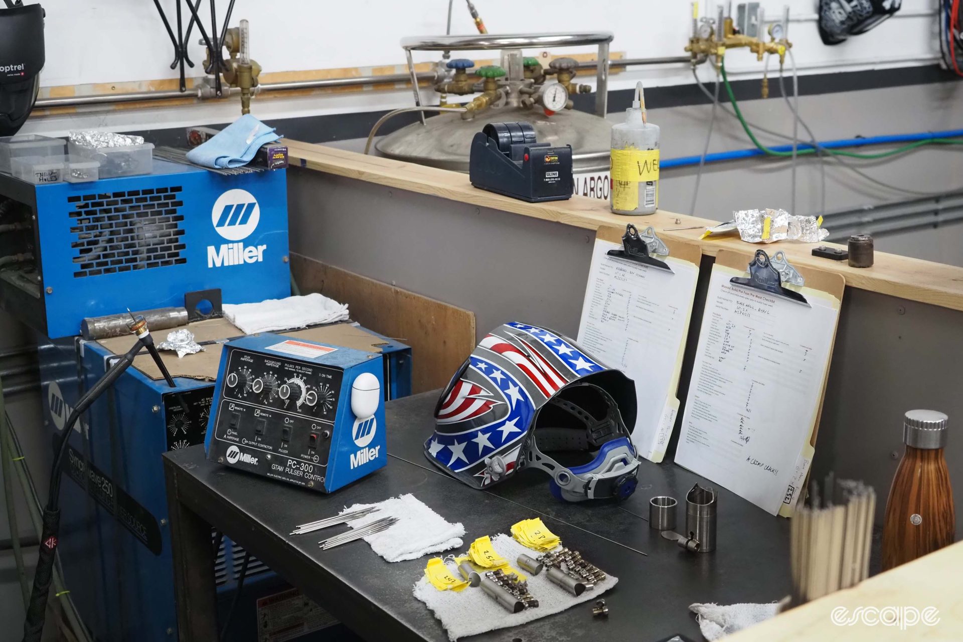 A neat and orderly welding station with a welding control module, mask, tungsten rod and small parts along with the order sheet.