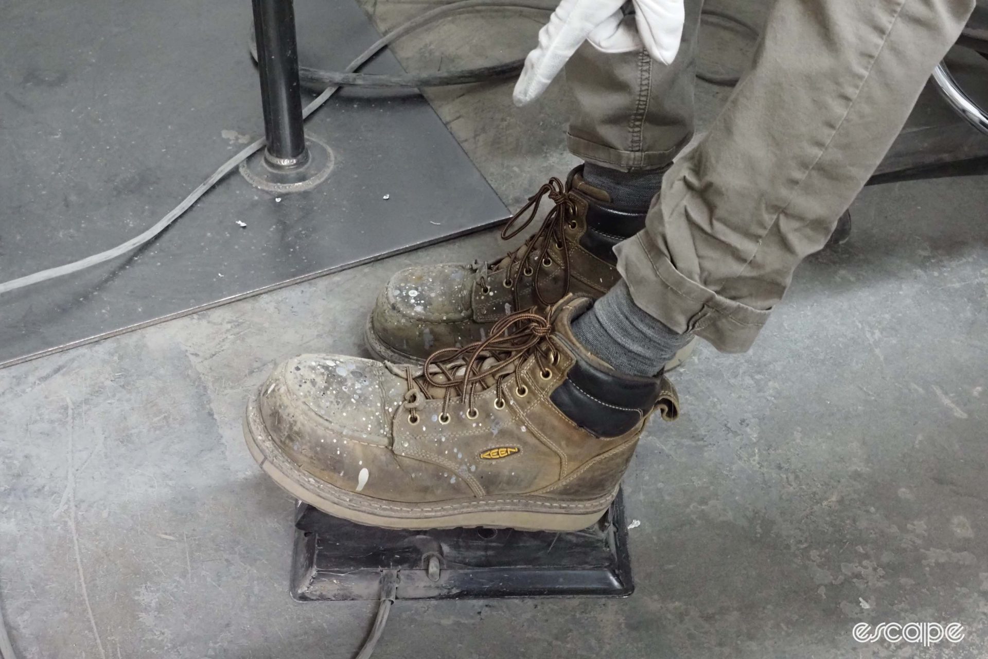 A welder in weathered Keen boots operates a welding foot pedal to control amperage. The boots are stained and dappled with paint and other substances.
