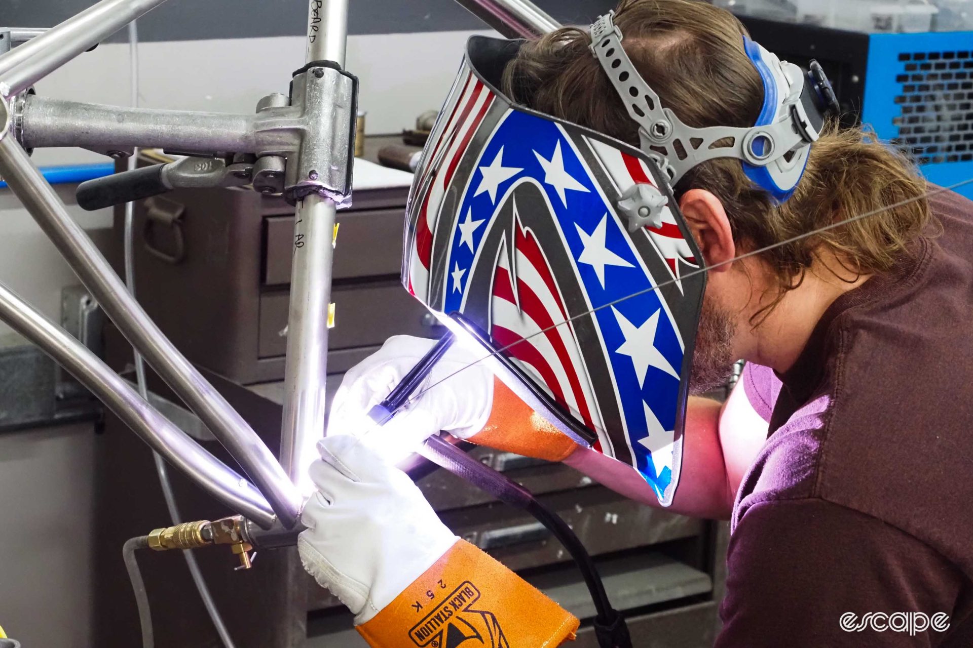 A welder in an American-flag stylized mask leans in to weld a bottom bracket juncture. His hands are brightly lit by the welding arc.