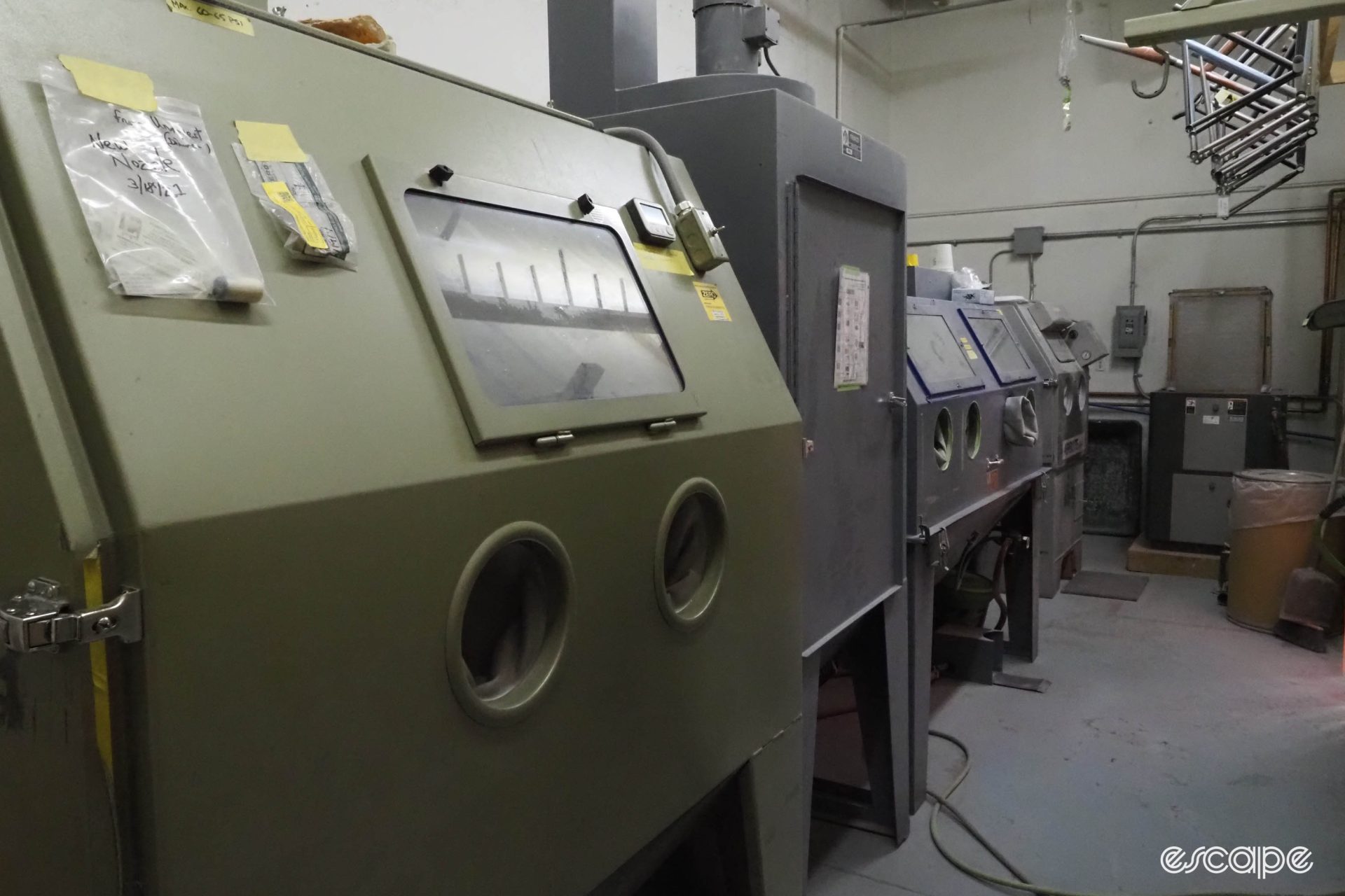 A large bead-blasting machine sits in front of several others. The olive-green machine has two holes for an operator to put their hands in to move the frame around inside.