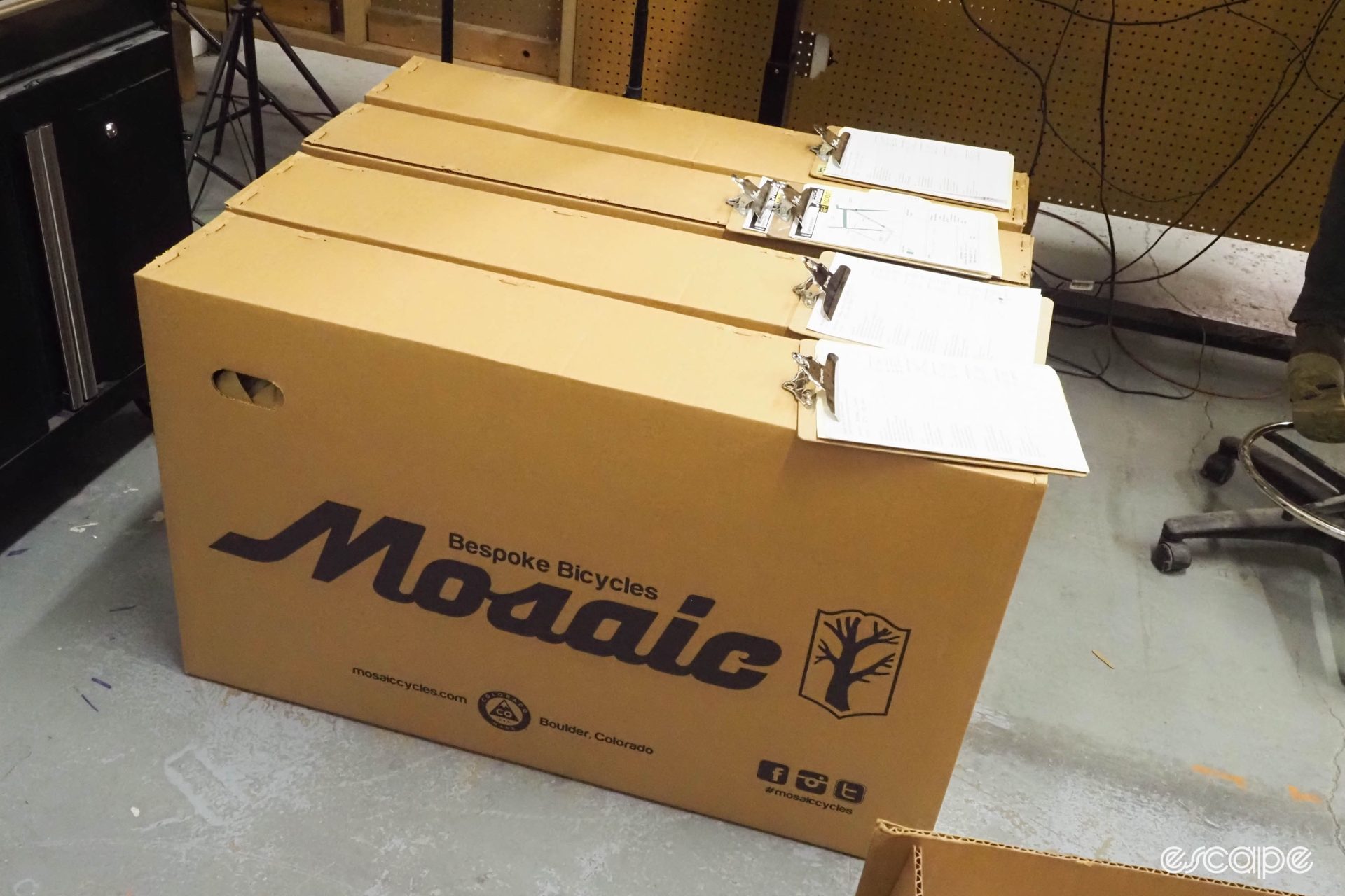 Four large shipping boxes marked "Mosaic Bespoke Bicycles" sit with customer order sheets.