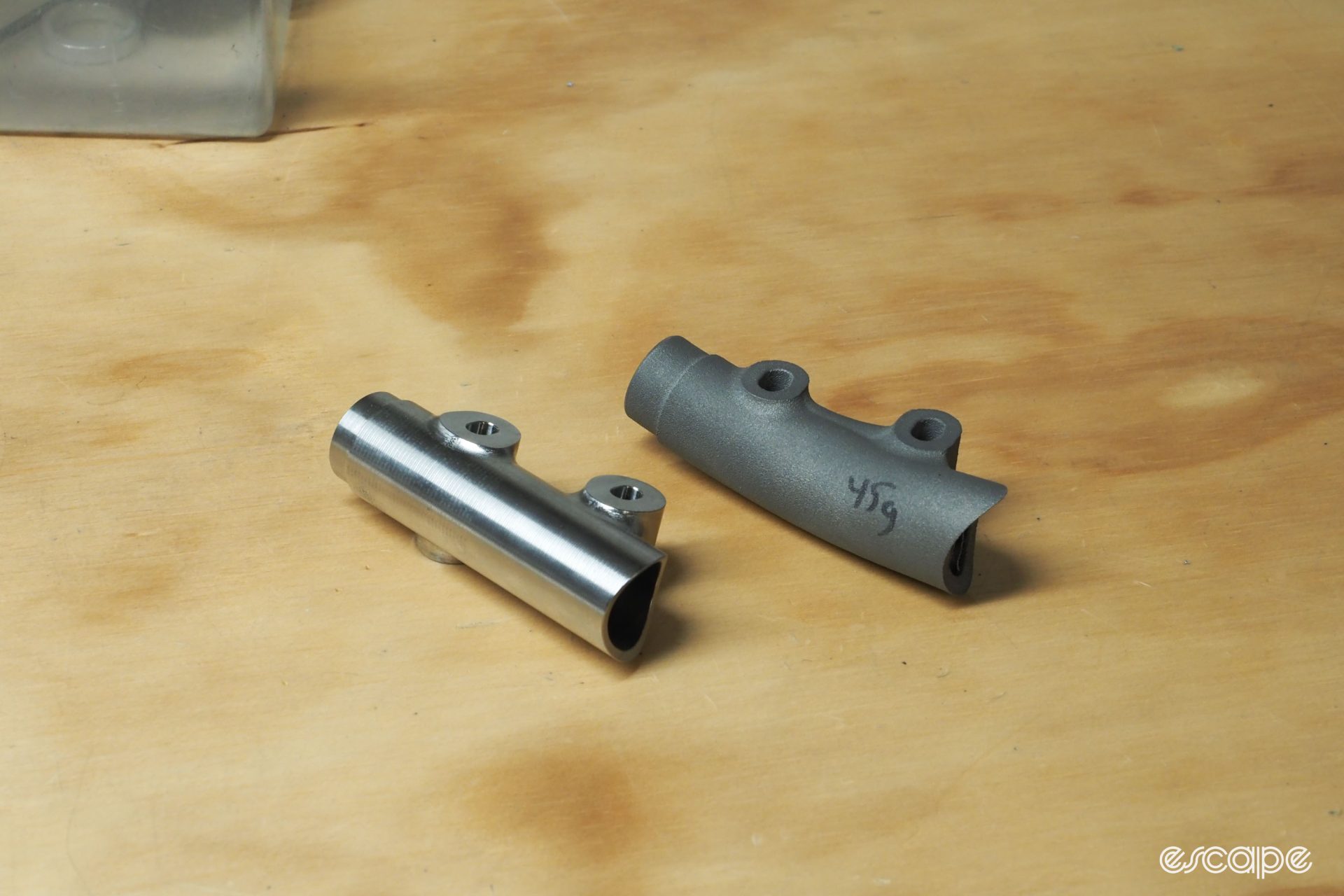 A conventionally made rear disc brake mount shown next to a 3D printed model. Both are titanium, but the conventional one has a distinctive satin finish while the 3D one is a rough, grey matte.