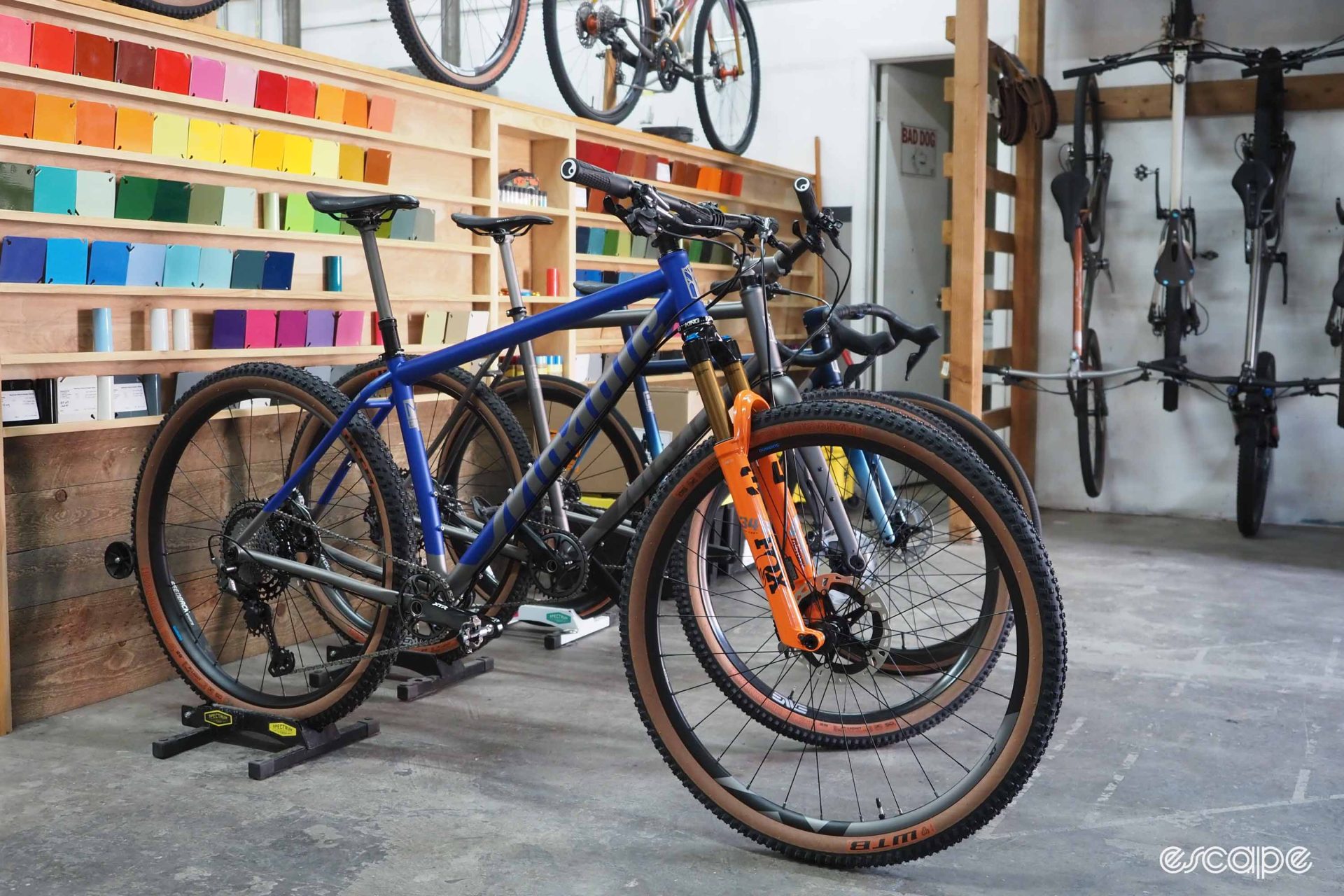 Several demo bikes sit on the Mosaic shop floor, including a hardtail mountain bike. Behind are rows of paint chips.