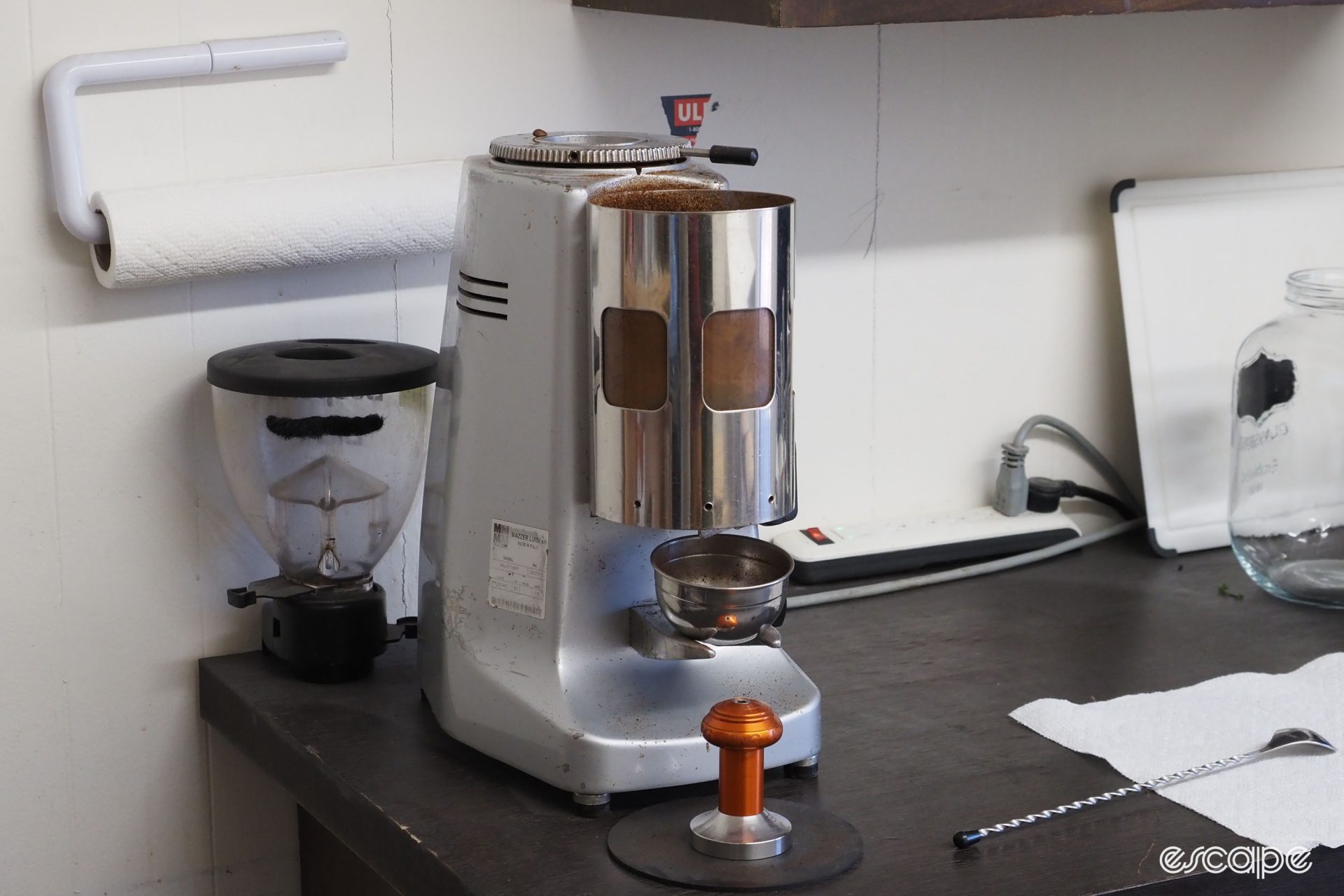 An old bean grinder for coffee sits on a bench, along with a Chris King espresso tamper in orange.
