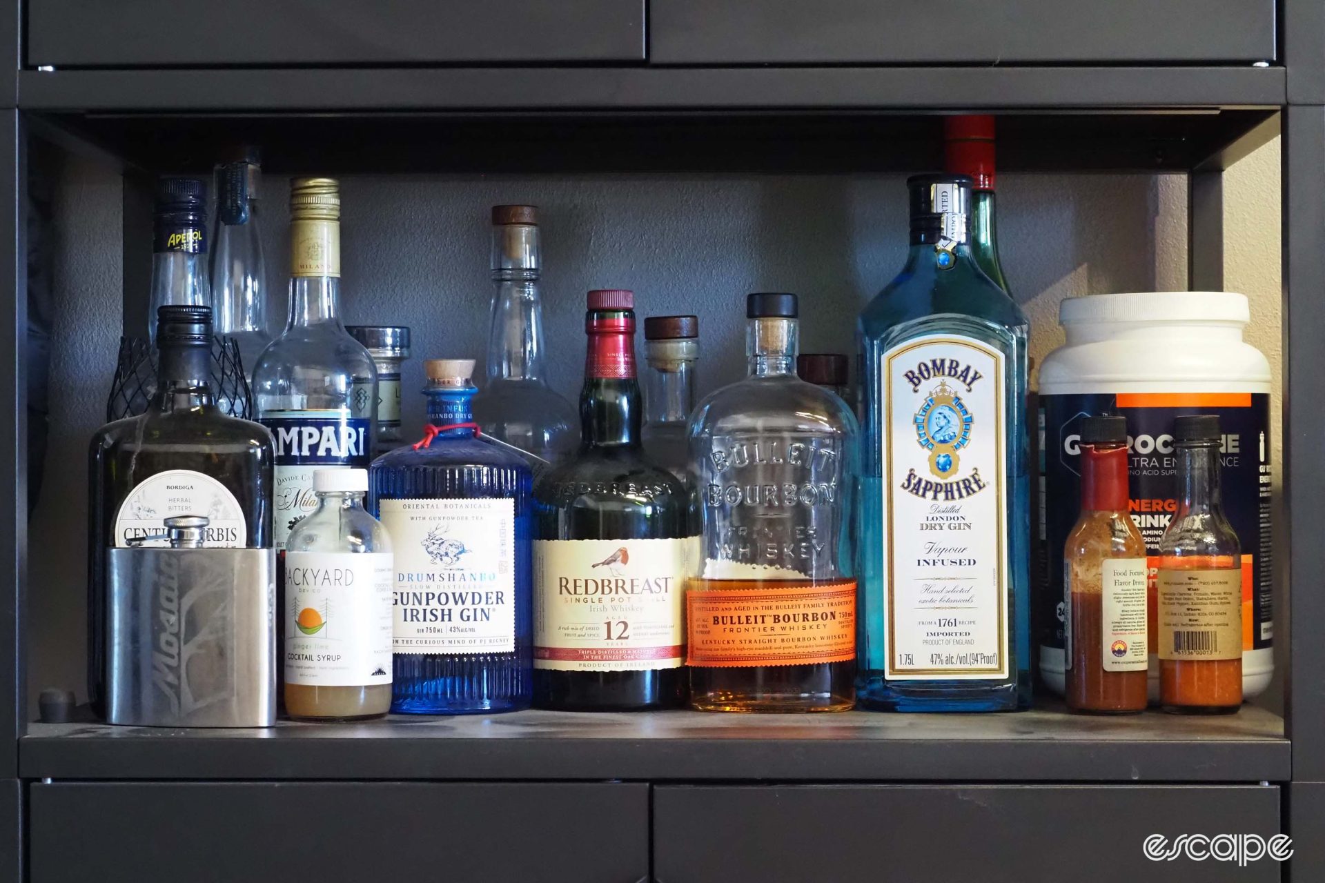 A row of liquors on a shelft, ranging from gin to bourbon, and a Mosaic-branded flask.