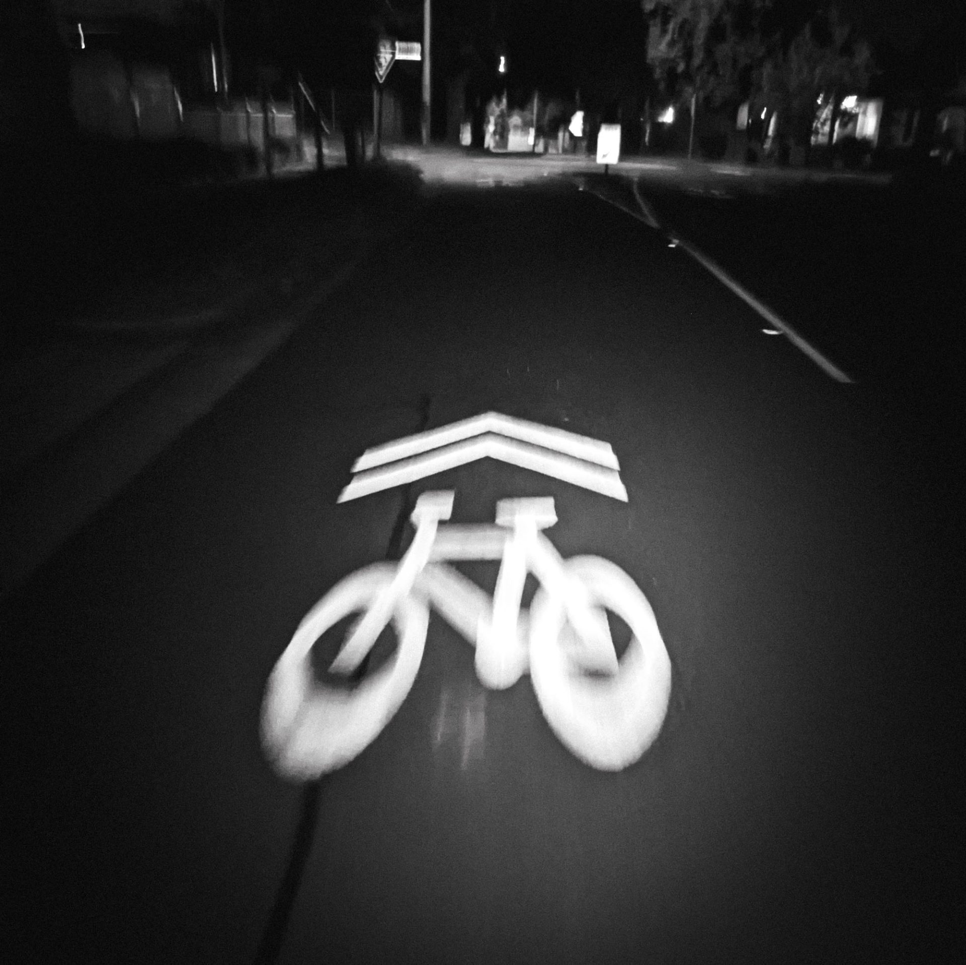 A moody, black-and-white night shot of a bike lane. The "Sharrow" signal of a bike and two arrows is slightly blurred but pops on the blackened pavement. Ahead, an intersection looms.