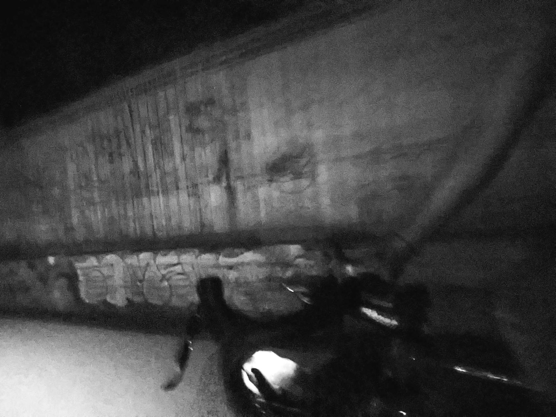 An almost impressionist black and white shot of a rider at night. A ghostly arm reaches down to a handlebar, silhouetted against a concrete wall, slightly blurred from motion.