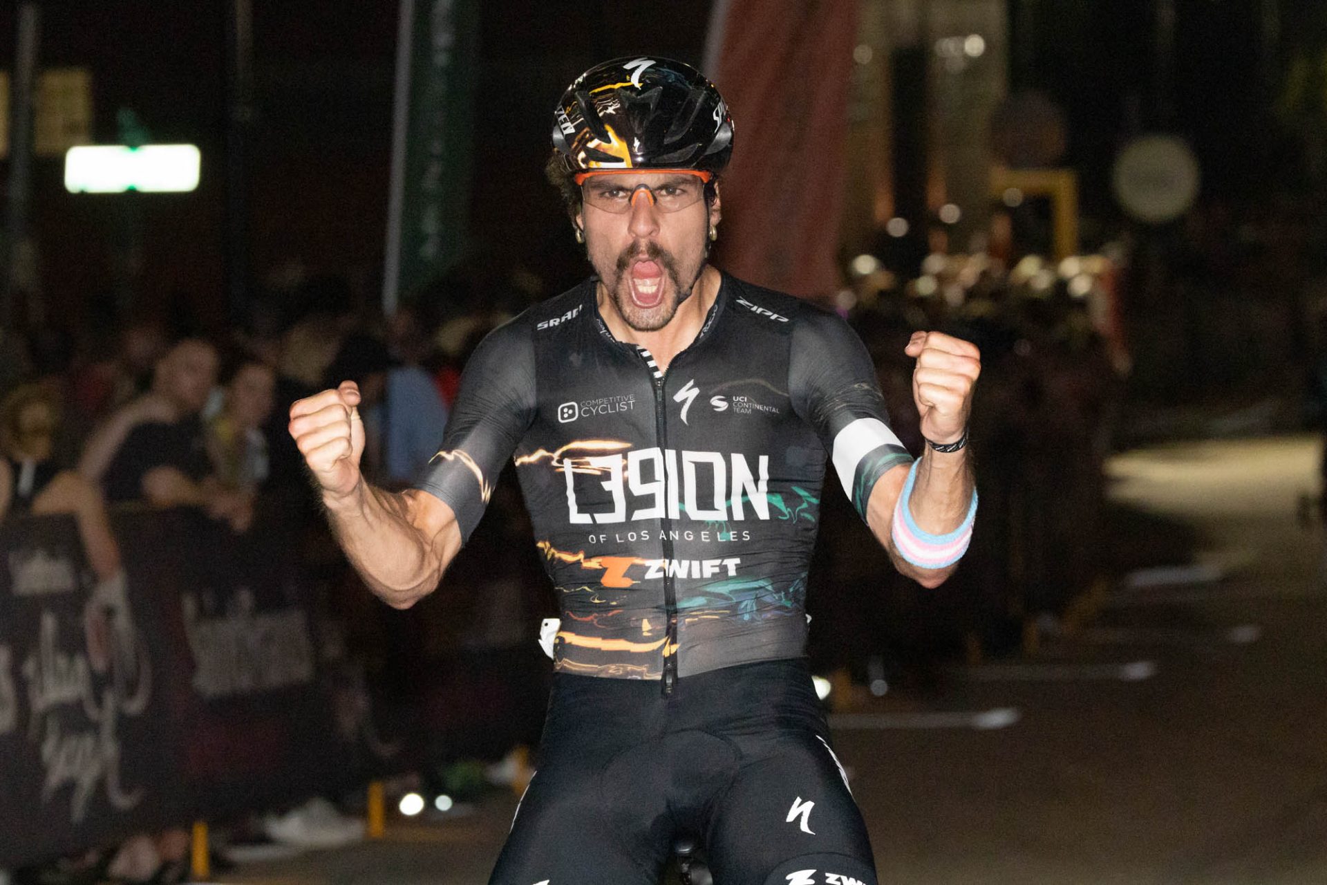 Sam Boardman celebrates a win. He's racing at night, in a black L39ION kit, and he's sitting up on the bike, both arms raised and hands clenched into fists as he lets out a yell.