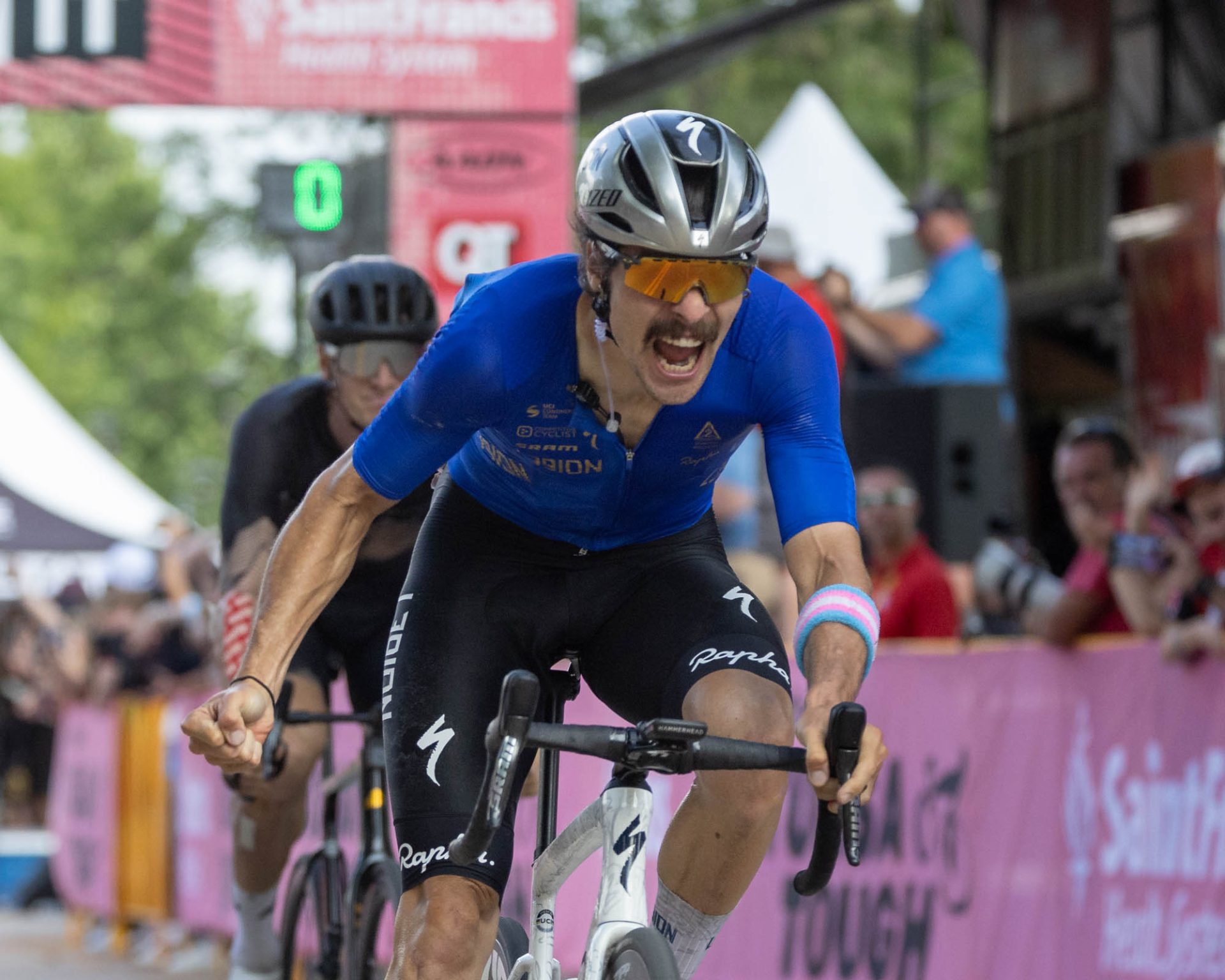Sam Boardman celebrates as he wins Tulsa Tough. It's immediately post-sprint and he has one hand off the bars, clenched in a fist, while his mouth is open in a celebratory yell. On his left forearm is the blue, pink, and white armband to signify support for trans rights.