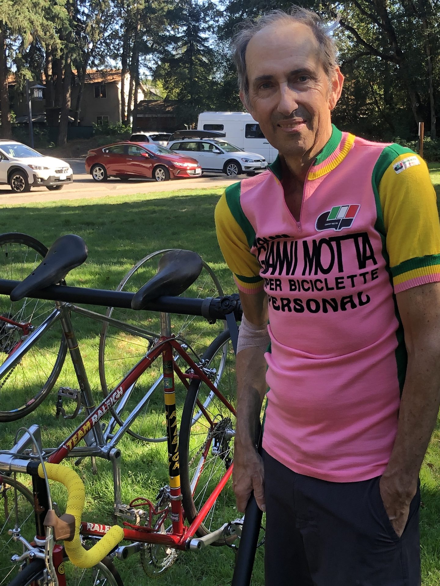 Tim Rutledge smiles as he poses with an old Raleigh road bike. He has grey, thinning hair and is partly bald, and wears a pink, green, and yellow Motta jersey like the ones designed for the Giro. He has a slight smile on his face.