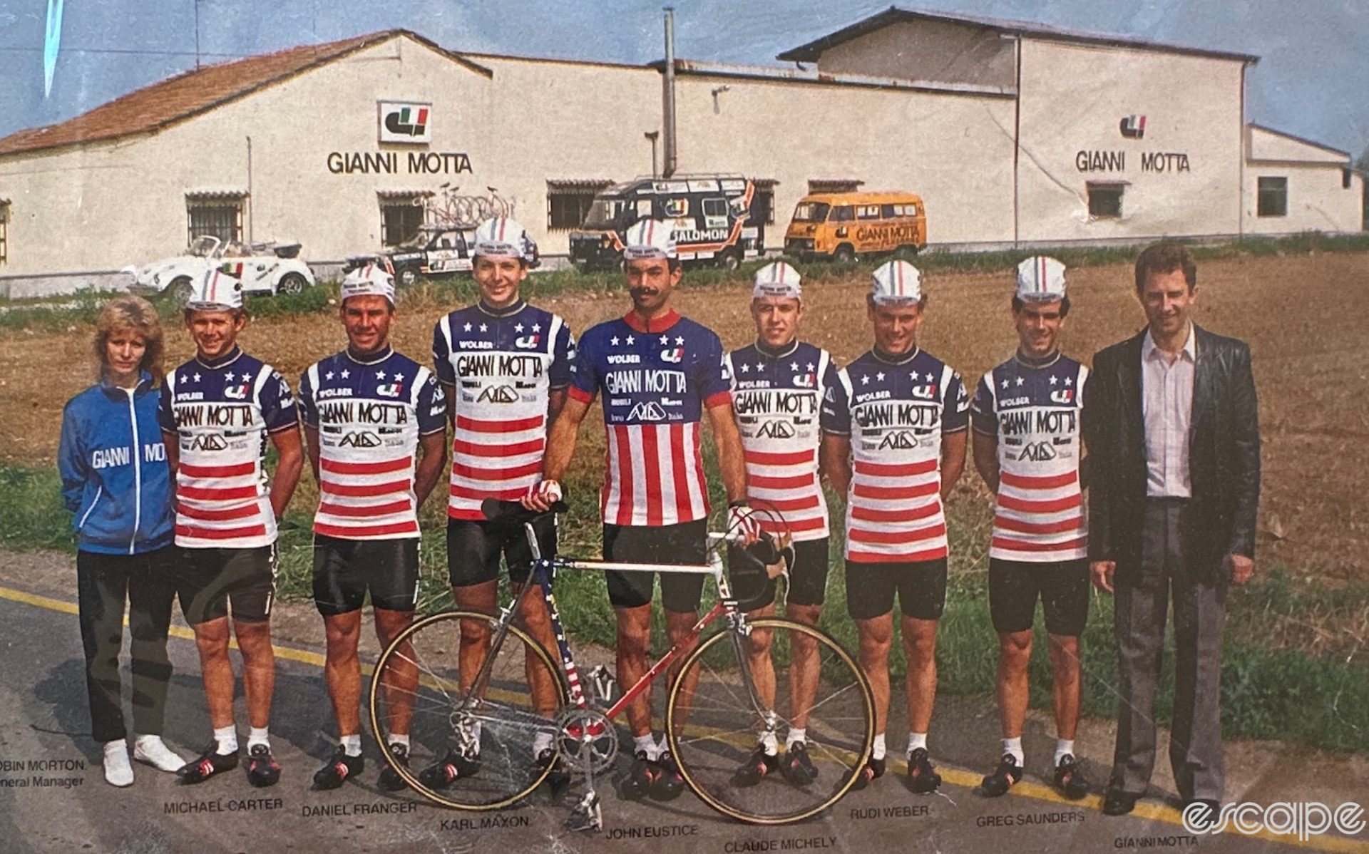 Members of the Gianni Motta team pose in front of the bike factory in Italy. There are eight riders, in the stars-and-stripes motif. John Eustice is in the center, flanked by Daniel Franger, Michael Carter, and director Robin Morton to his right, and Claude Michely, Rudi Weber, Greg Saunders, and Motta to his left. Morton is wearing a blue Gianni Motta trainer and black pants, and Motta is in a black leather blazer. A Motta bicycle stands in front of the team.