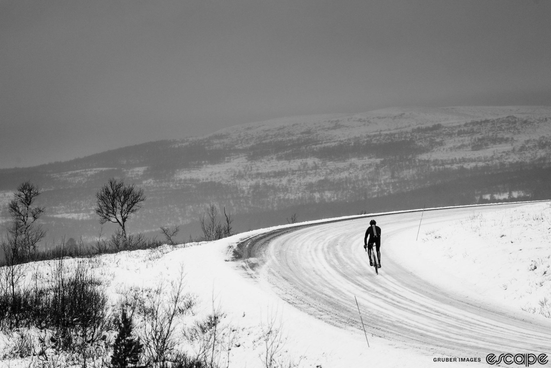A solo rider climbs a snow-covered road around a curve. A broad, gentle hill rises in the background, the forested ridge covered in snow. The light is flat and dark.