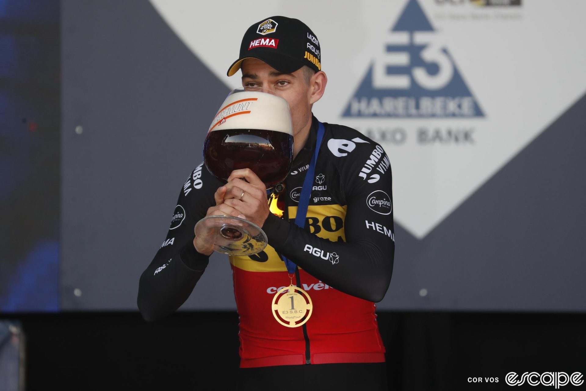Wout van Aert drinks from a massively oversized beer on the podium of the 2022 E3 Saxo Classic.