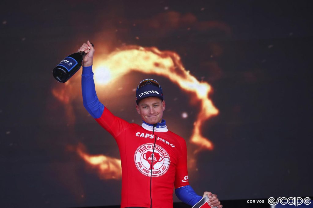 Mathieu van der Poel salutes the crowd on the podium of the E3 Saxo Classic. He holds a bottle of champagne while a pyrotechnic display lights behind him.