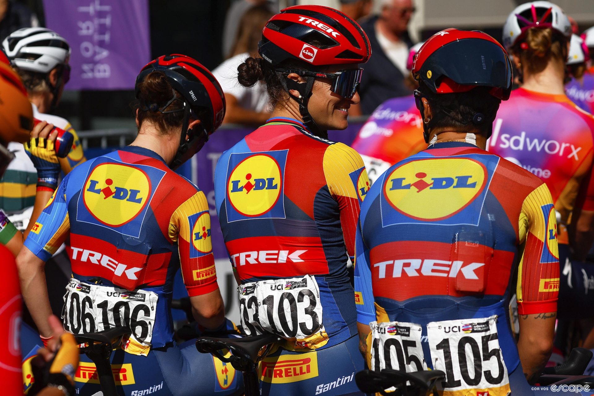 Three Lidl-Trek riders laugh together while waiting for a race to start. The back of the jerseys show the color blocking, with a yellow Lidl circle surrounded by slanted blocks of blues and red, a red band around the center with the Trek logo, and a blue lower third.