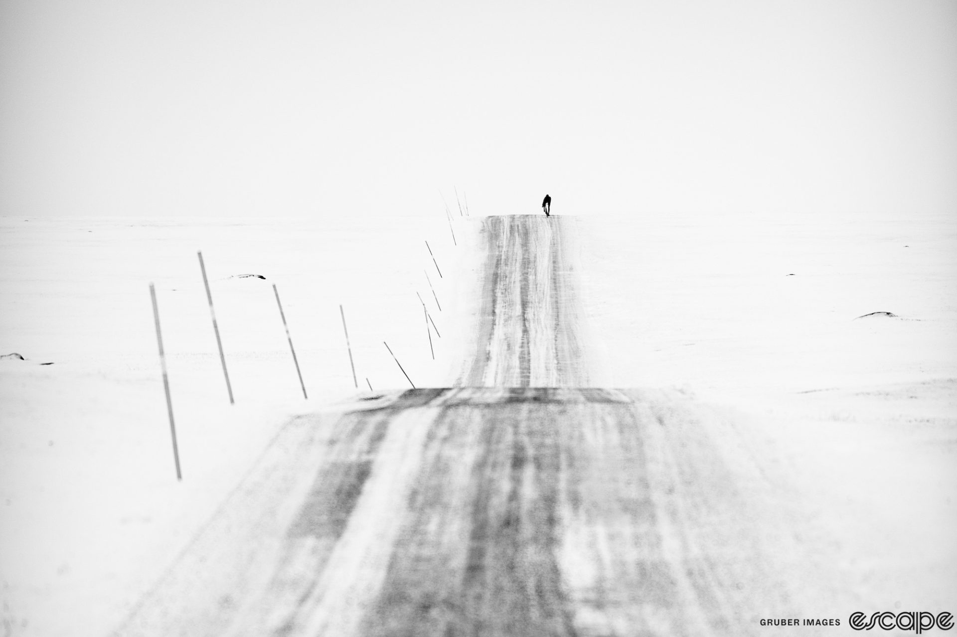 A lone rider is seen in the distance cresting a roller on a snowy road. The road is covered in snow but plowed, and the surrounding landscape is entirely winter white, with flat light creating an almost black-and-white effect.