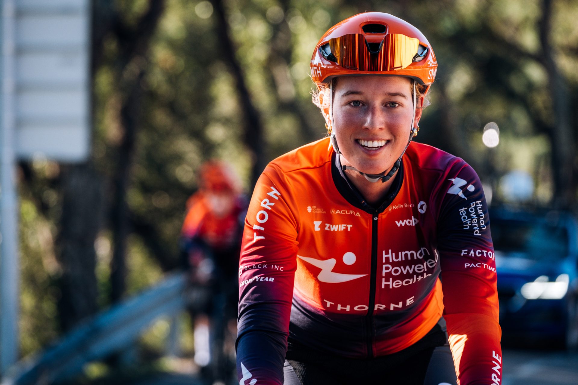 Alice Barnes smiles at the camera while riding up a climb in Spain. The HPH kit is purple to orange fade, with alternating colors on each shoulder and an orange body with a purple trim at sleeves and lower right hem. The purple describes a kind of diagonal theme across from left shoulder to right hip.