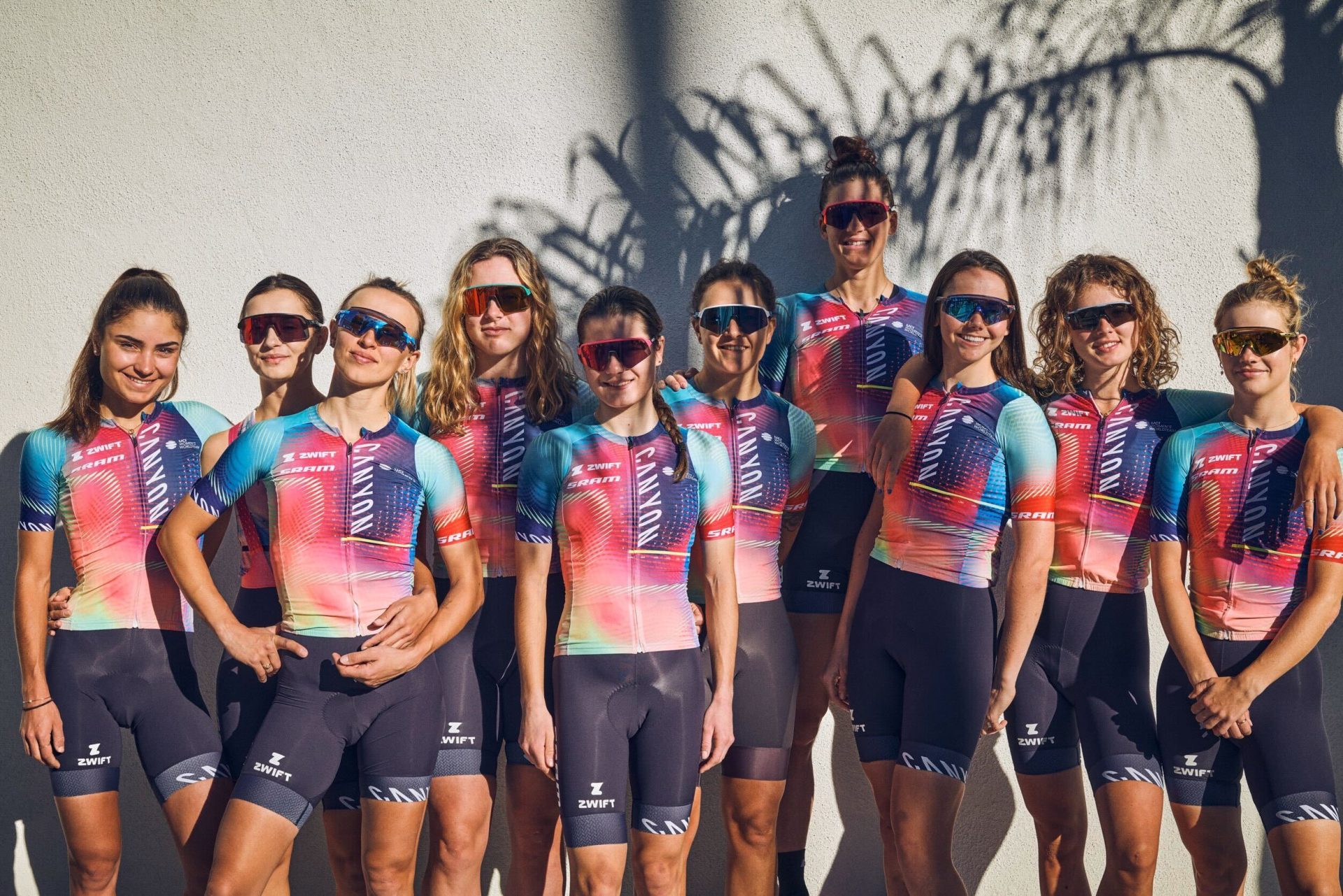 The Canyon-Sram team models their new kit outside a coffee shop in California. The jerseys feature afade design with light blue shoulders set against a dark blue to pink fade across the kit, ending in a greenish yellow at the lower right hem.