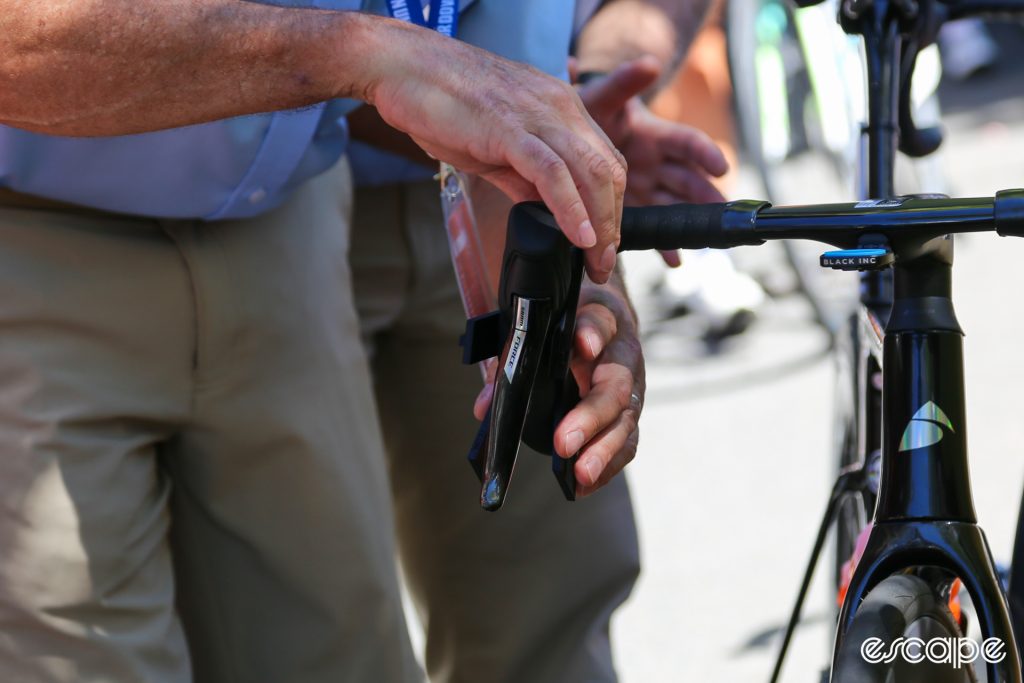 The image shows a UCI commissaire checking the inward rotation of a brake lever