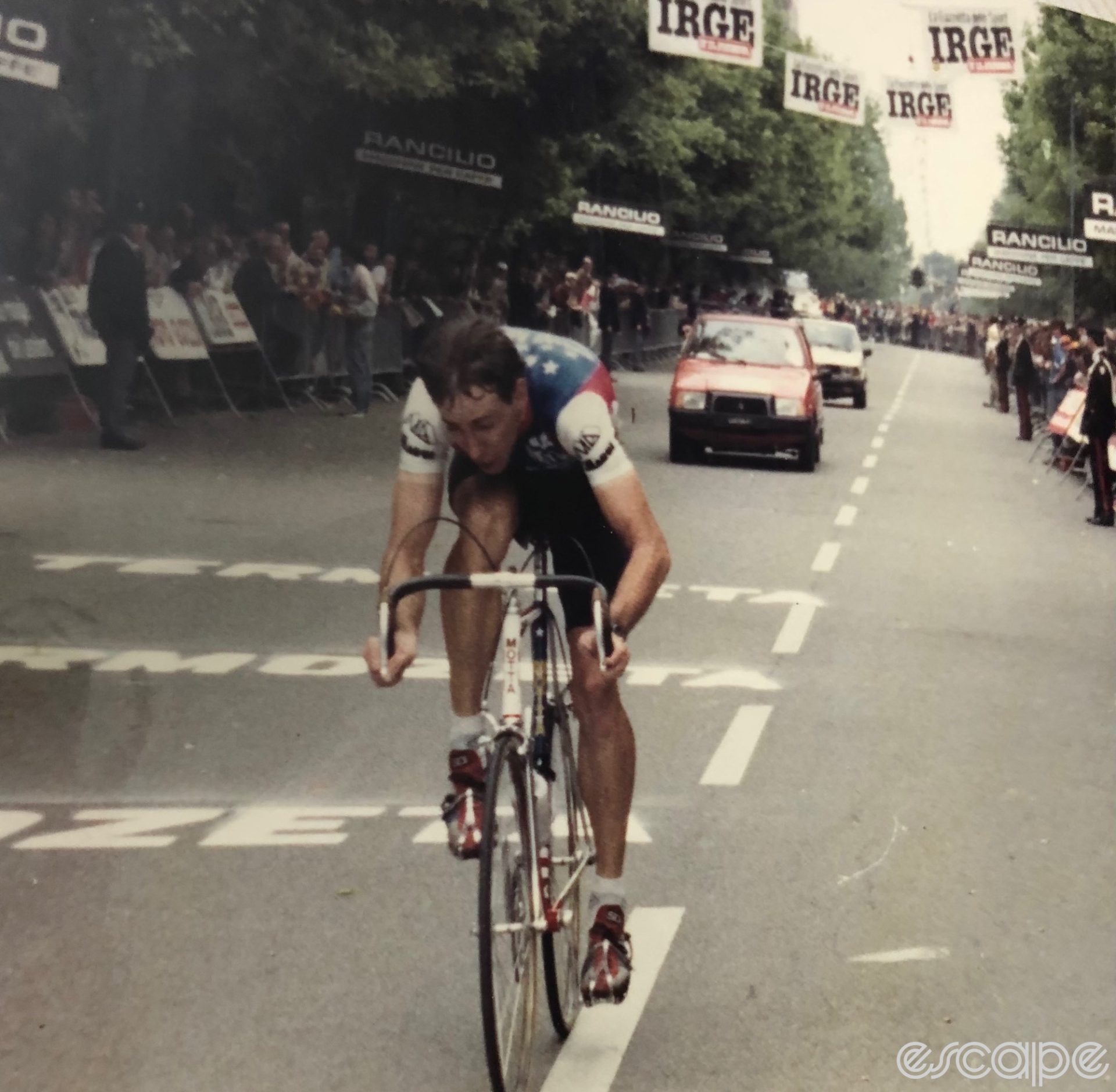 Tim Rutledge races a time trial at the 1984 Giro. He's alone on the road, with fans crowded behind barriers and two cars following behind. There is a look of intense suffering on his face.