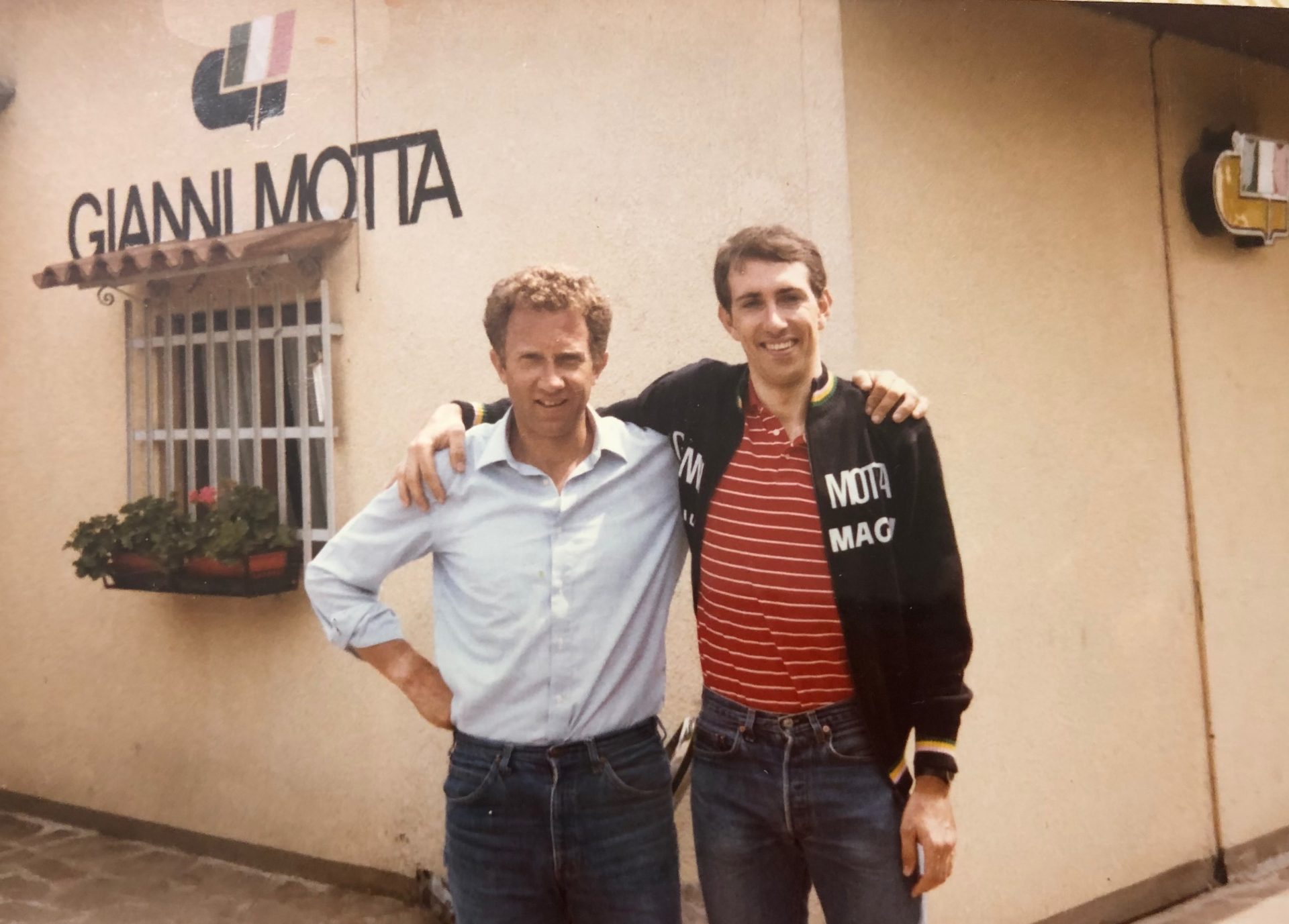 Tim Rutledge stands arm in arm with Gianni Motta for a picture outside the Motta factory. Rutledge is tall and boyish-looking, and is wearing a Motta-branded trainer jacket and jeans and has a wide smile. Motta is slightly shorter, with a confident bearing and slight smile, dressed in a light blue button down and jeans.