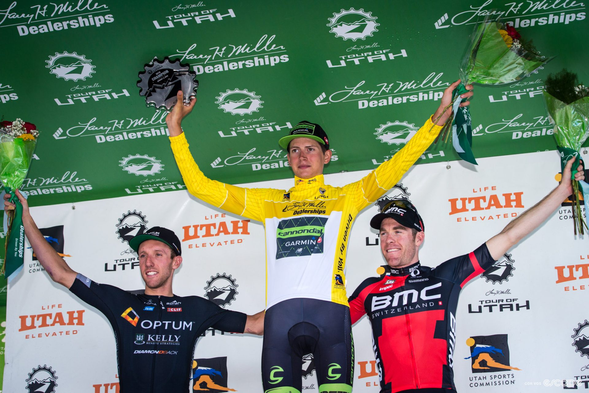 Joe Dombrowski celebrates on the podium of the Tour of Utah. He's in the yellow jersey of race leader, and is flanked by Michael Woods on his right and Brent Bookwalter to his left.