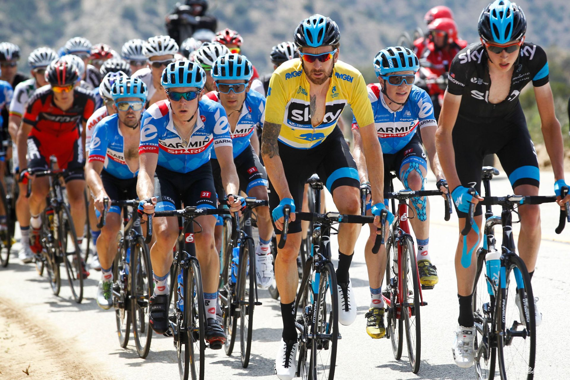 Joe Dombrowski leads a line of riders at the 2014 Tour of California. Yellow jersey and teammate Bradley Wiggings is next to/slightly behind him, with a pack of blue-jerseyed Garmin riders tucked in right behind.