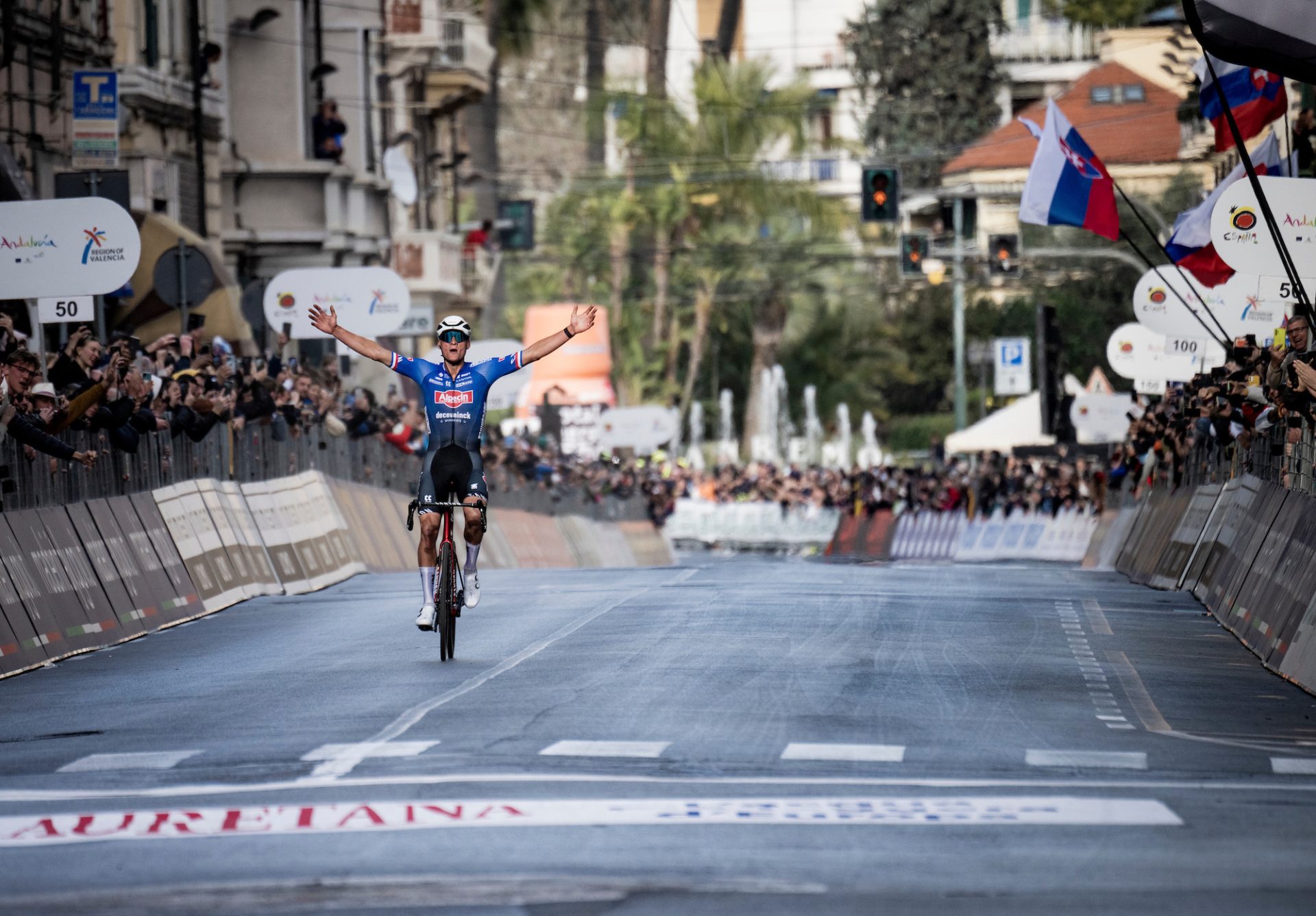 Mathieu van der Poel holds his arms aloft in a wide victory salute in Milan-San Remo. He's on the famous Via Roma, and behind him the fan-lined barriers frame an empty road as he rolls to the finish line solo.