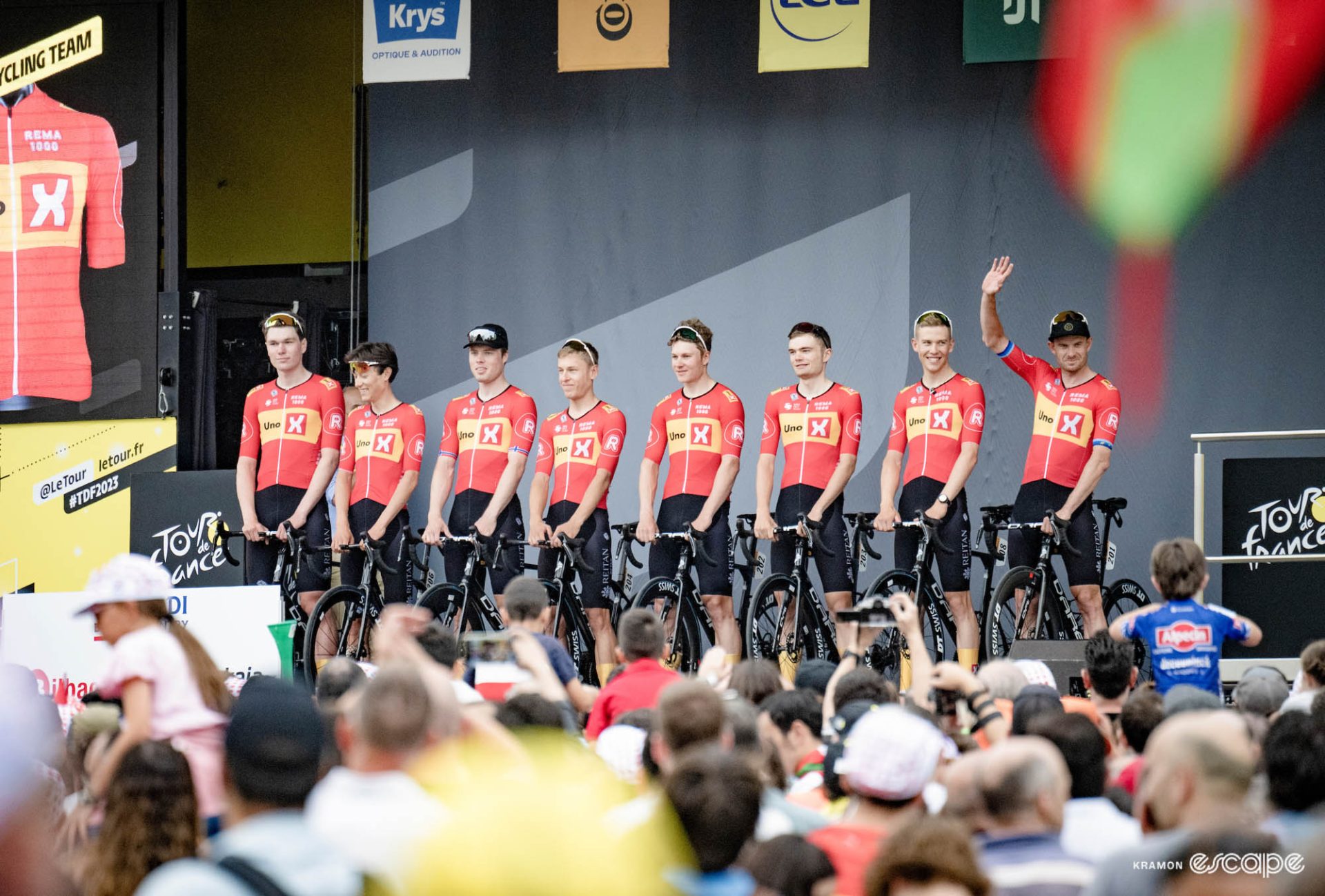 Alexander Kristoff stands on a stage along with his Uno-X teammates at the 2023 TdF team presentation. He waves to the large assembled crowd as his seven teammates stand next to him in their kits and team bikes.