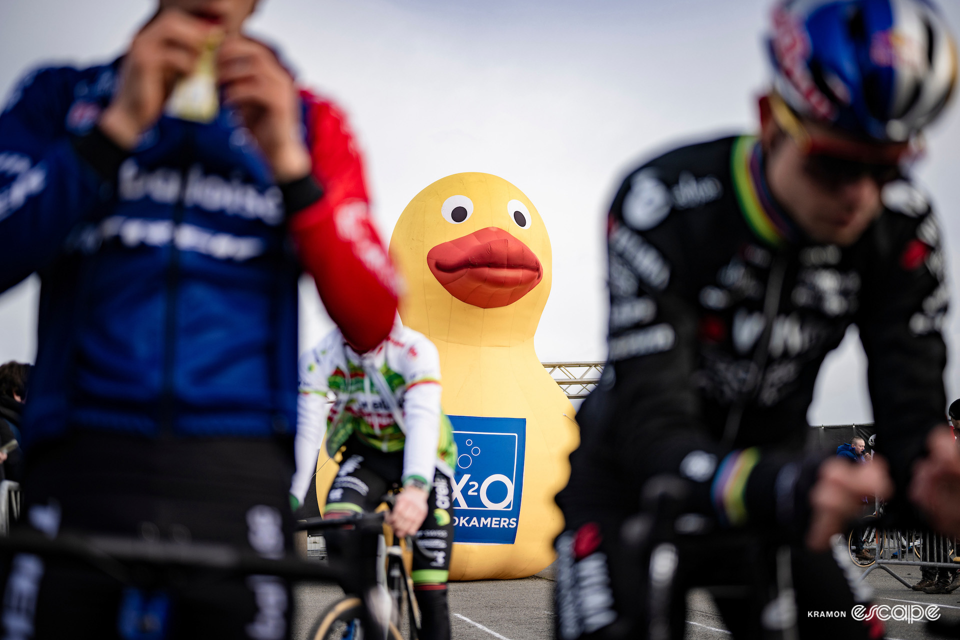 The large inflatable duck that announces the X20 Badkamers sponsor behind the start line at X2O Trofee Koksijde - Vlaamse Duinencross, Wout van Aert and Thibau Nys out of focus in the foreground.