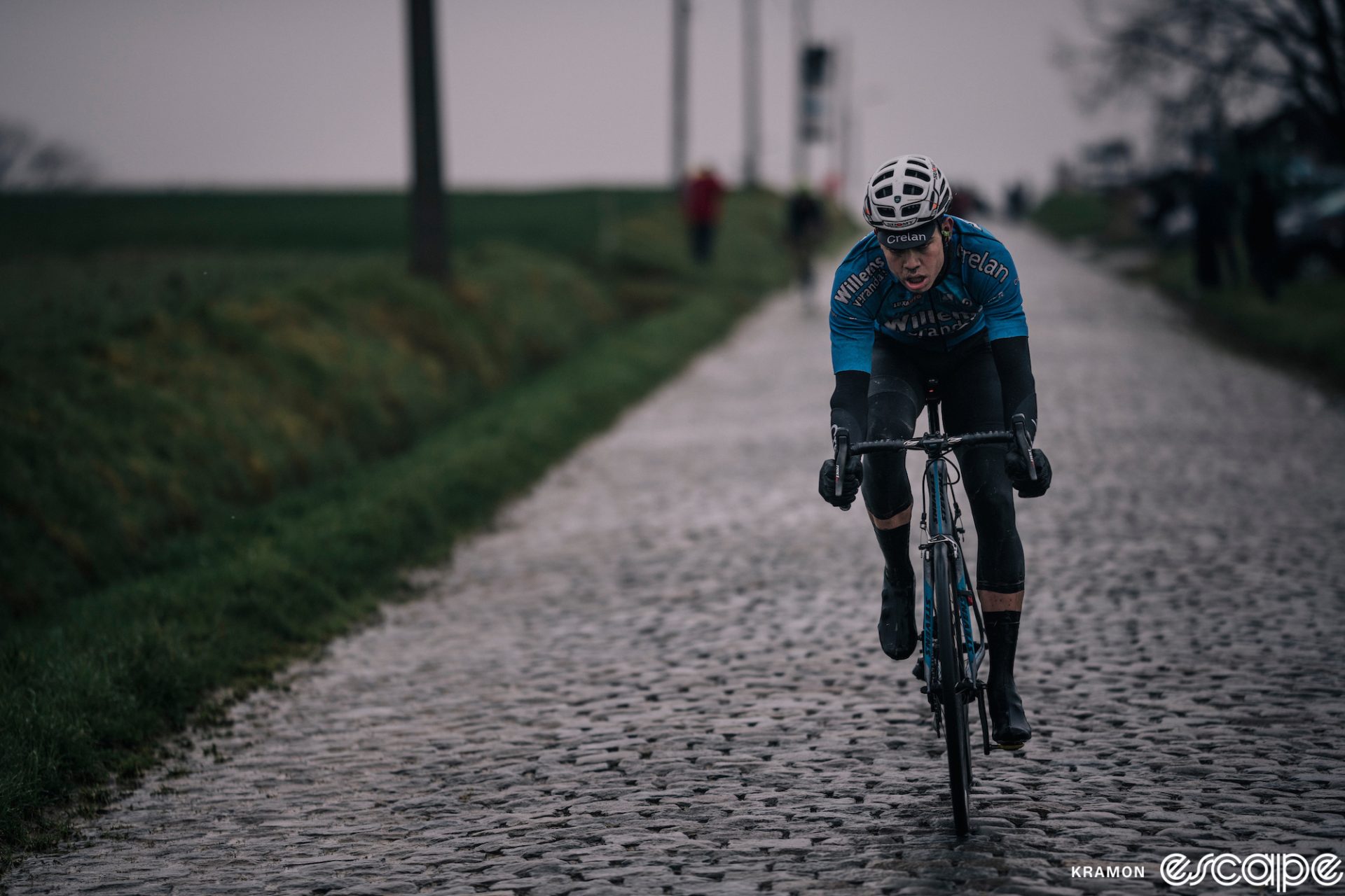 Wout van Aert rides on cobblestones at the Dwars Door Vlaanderen race in 2018. It's cloudy and rainy and the lighting is dark and moody. He's in the blue of Verandas Willems, a team he would soon force his exit from.