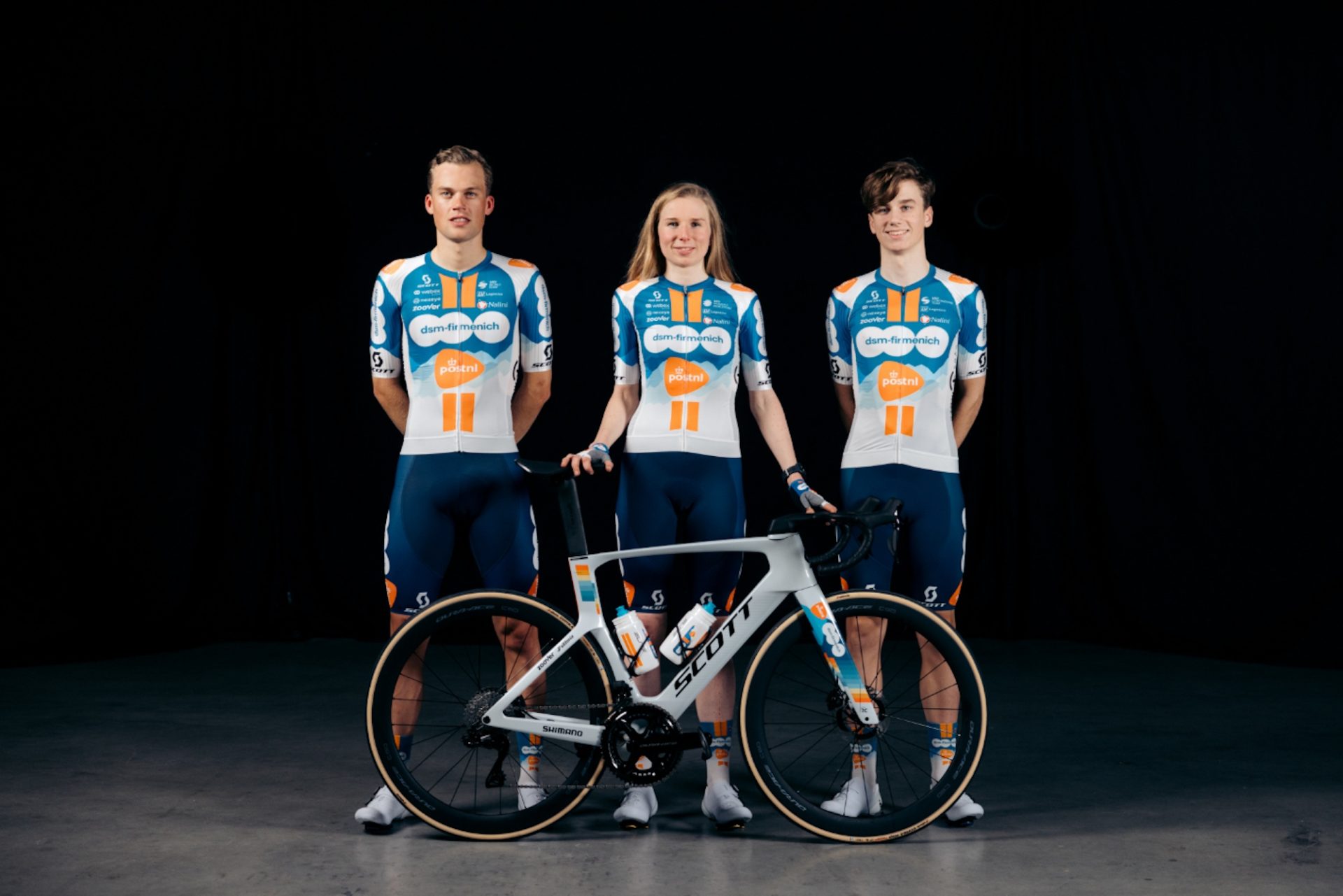 DSM Firmenich-PostNL riders display their new kit and bikes. Two male on either side of one female. The jerseys are white with a medium-blue block on the upper chest, a double orange stripe down the middle broken up by DSM and PostNL logos in white and orange, and a white lower half.