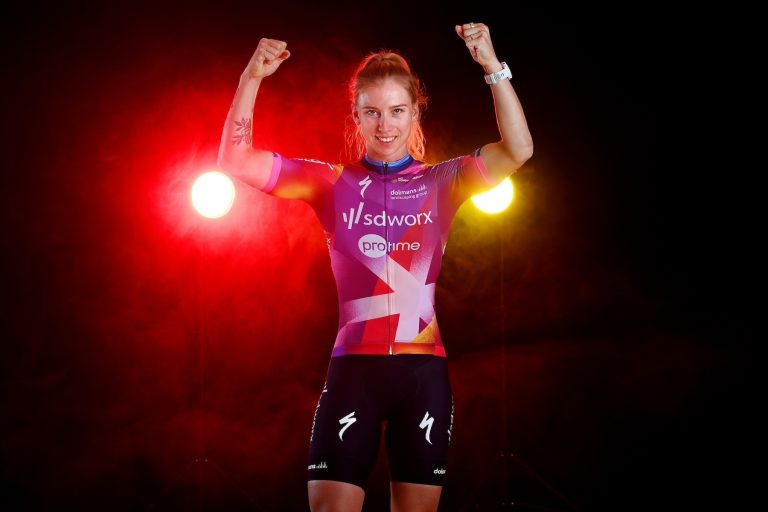 Lorena Wiebes raises her arms above her head wearing SD Worx new kit backlit by red and yellow lights.