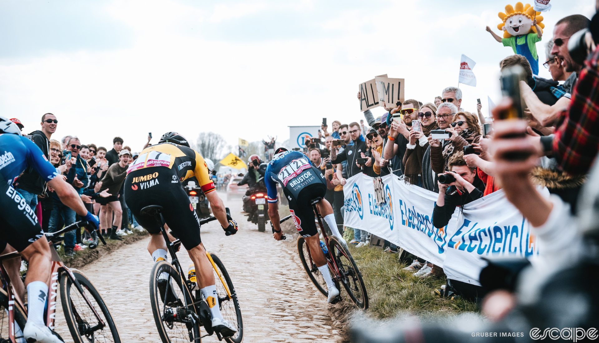 Mathieu van der Poel take a wide line through a corner at Paris-Roubaix. He's gone into the grass on the far side and is twisting his bike to get back on the cobbles, while Wout van Aert and Jasper Phillipsen behind take more conventional lines through the corner.