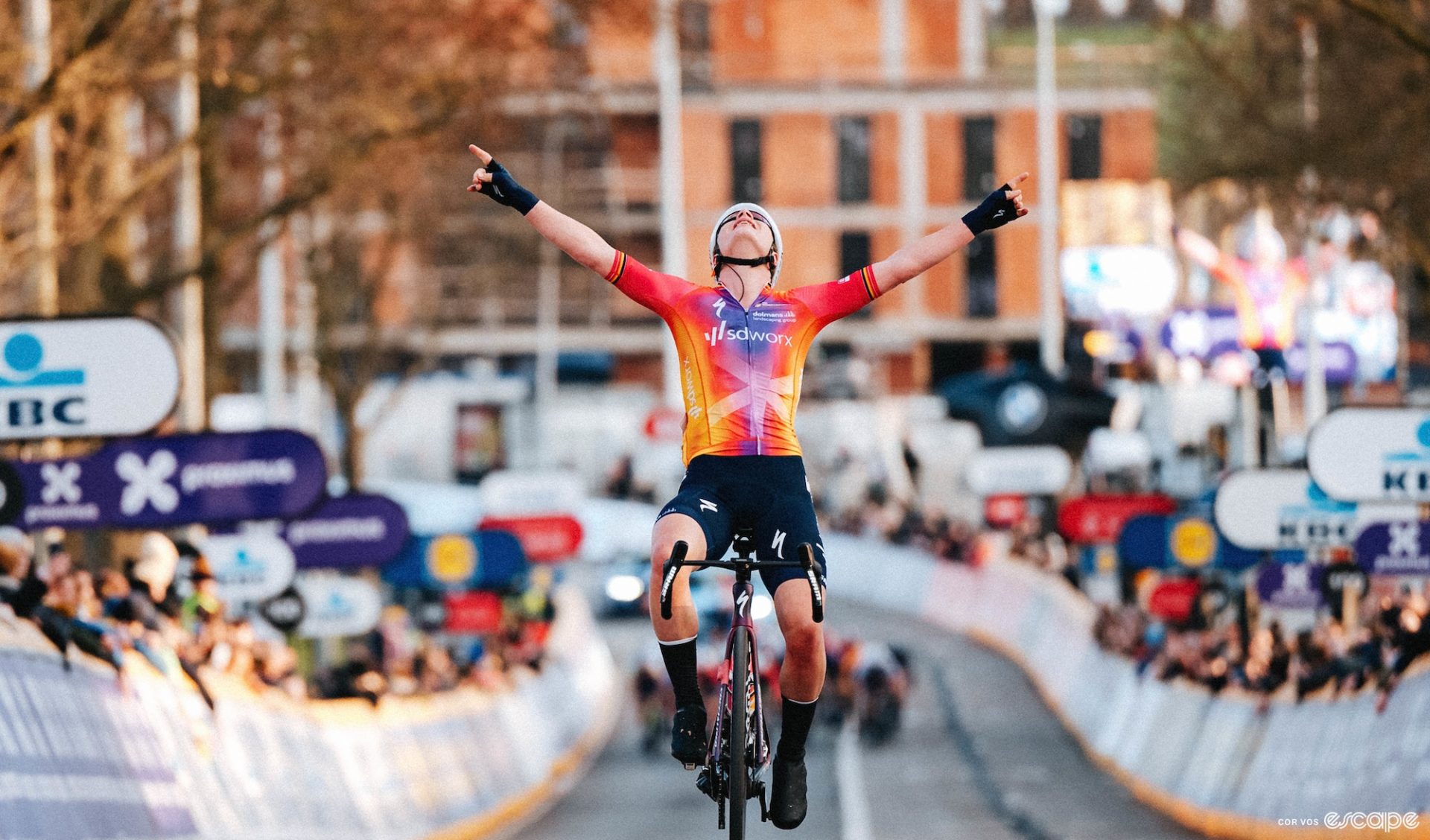 Kopecky raises both her arms in victory, with a group of riders sprinting in the background.