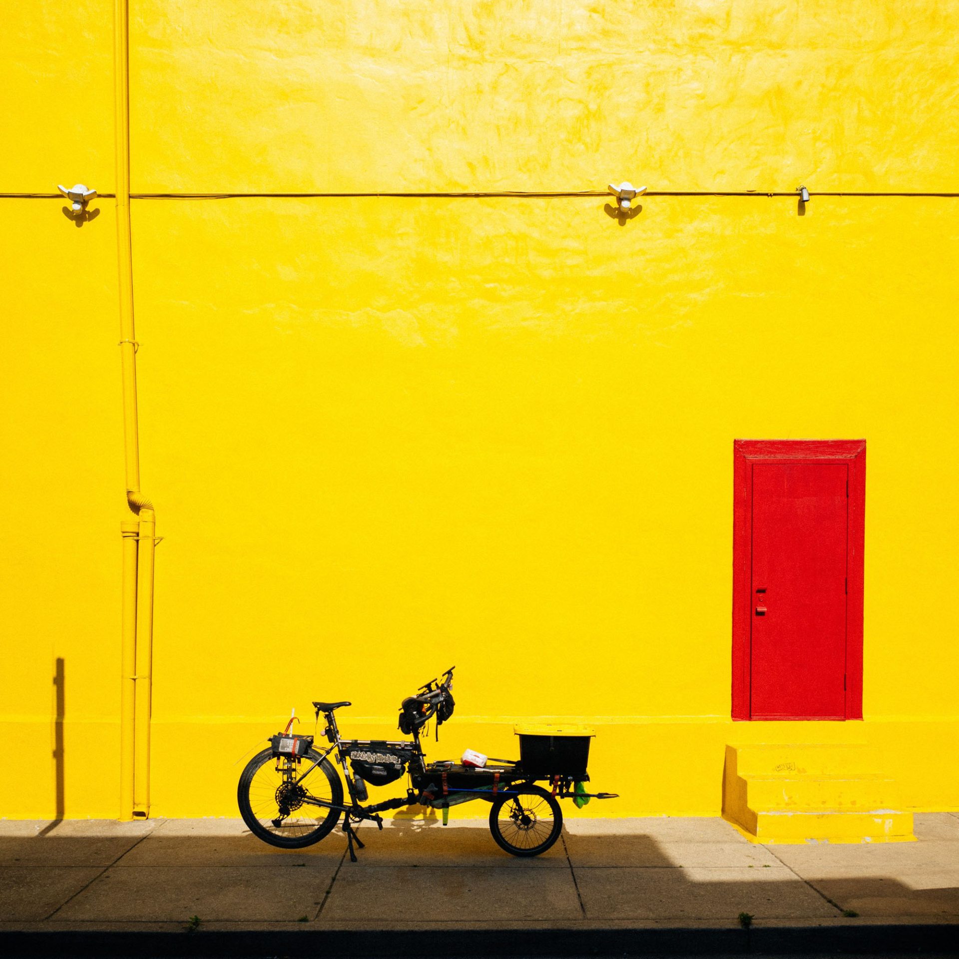 Binggeser's cargo bike in front of a brightly lit yellow building with a brilliant red door. It has a very abstract-art feel.
