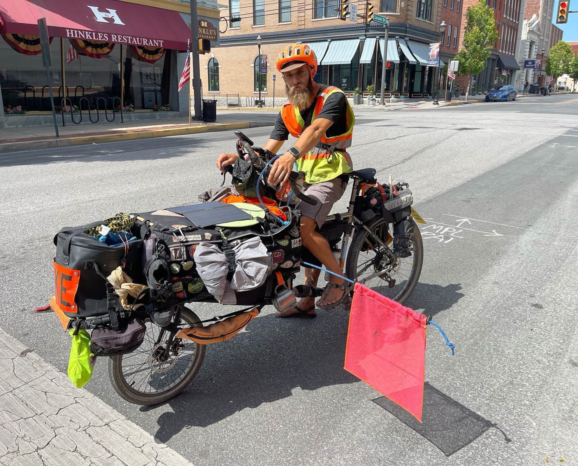 A bearded Binggeser on his fully loaded cargo bike. He has an orange helment, orange and yellow safety vest, and a large red flag protruding from the left side of the bike.