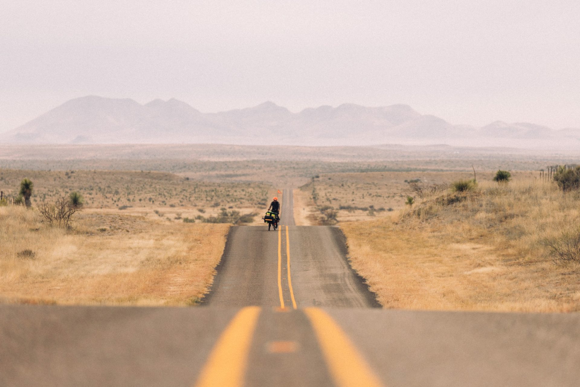 Binggeser rides his cargo bike on a lonely road in the desert. The two-lane tarmac stretches away into the distance over small rollers, with mountains in the background. There is no sign of human life except Erik.