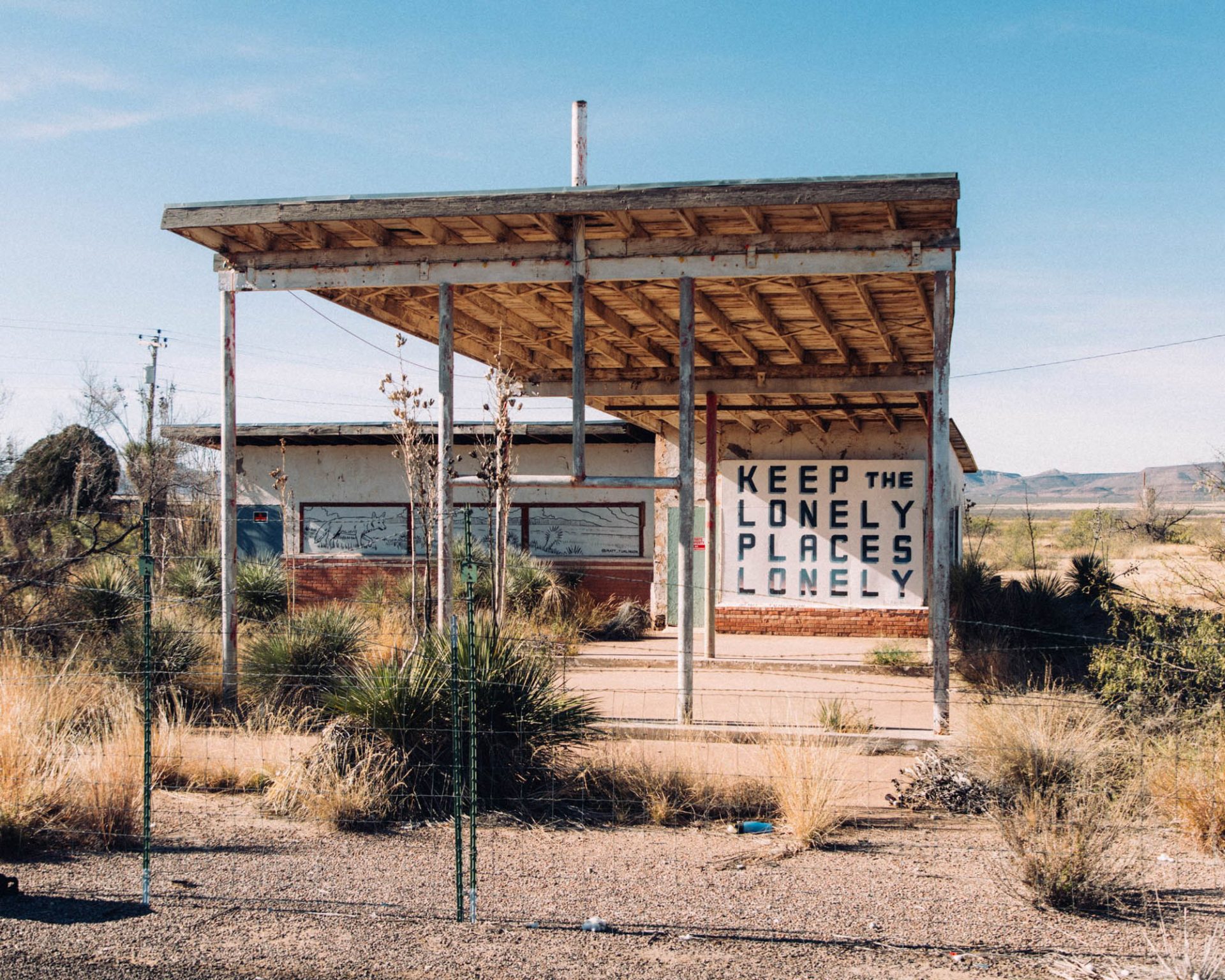 An abandoned gas station in the desert. The pumps are gone and the building is boarded up, with a large sign that reads "Keep the lonely places lonely."
