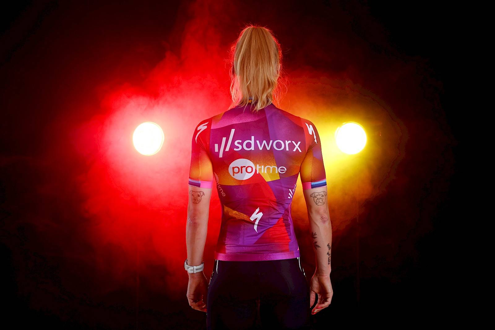 Lorena Wiebes stands facing away from the camera, the back of SD Worx's new jersey on display.  It's similar to last season, a blend of fuchsia, red, orange and yellow in both color blocking and fade.