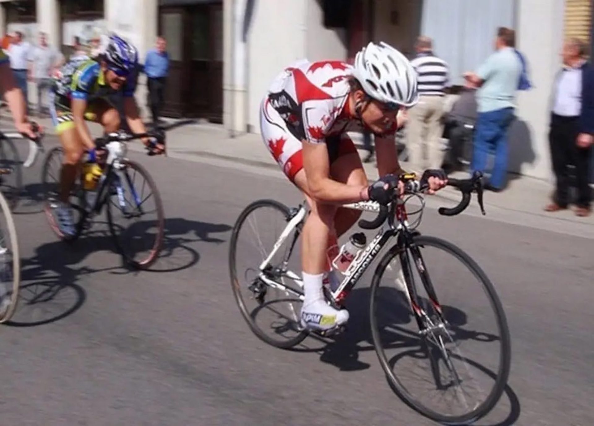 Keir Plaice races on a busy urban circuit. He's slightly blurred from the speed, as he pushes the pace bent low over his white and black Argon 18 road bike, dressed in the white and red maple leaf Canada national team kit.